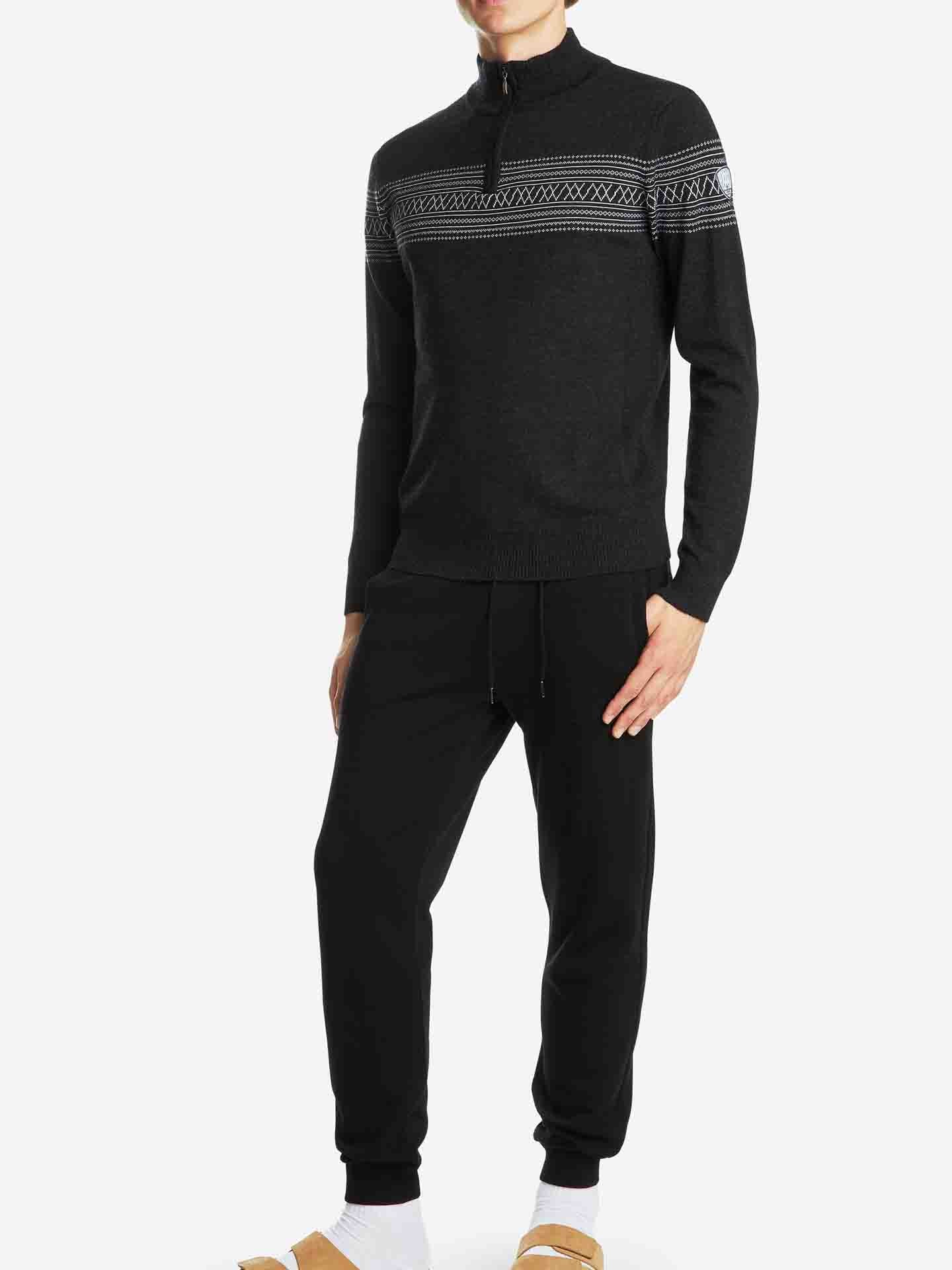 A person wearing a Signature Zip Up Men sweater by We Norwegians, characterized by black merino wool with white geometric patterns across the chest and a half-zip neckline, stands against a white background. They are also sporting black jogger pants paired with tan slippers and white socks.