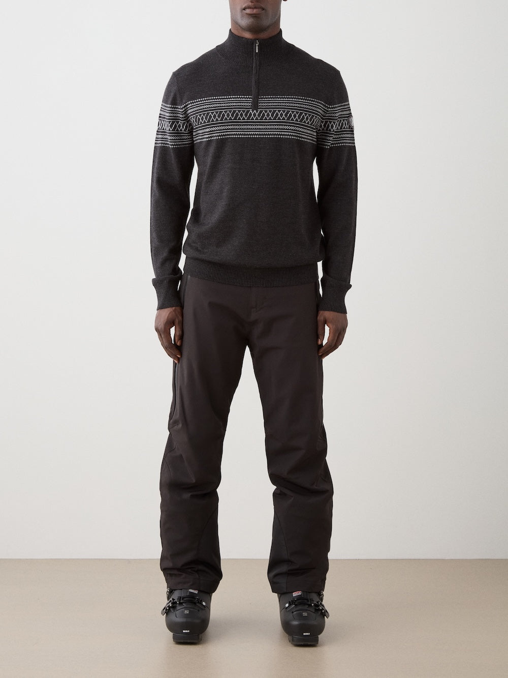 A person stands facing forward wearing the We Norwegians Signature Zip Up Men—a dark merino wool sweater with a white Norwegian Setesdal pattern—paired with black pants and black ski boots against a plain background.