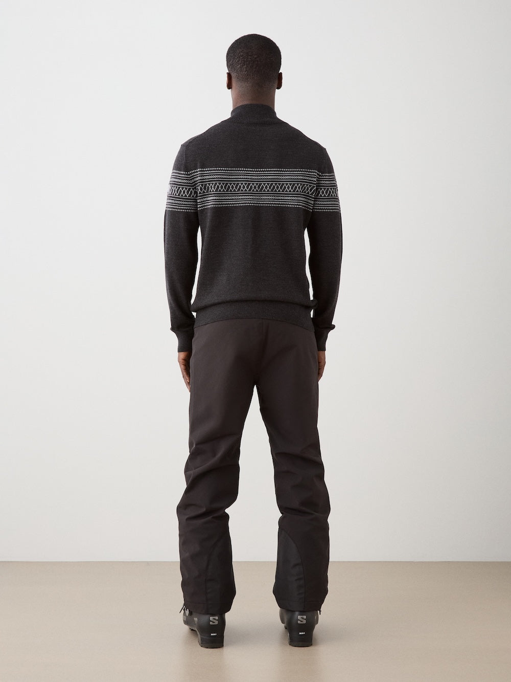 A person stands facing away, wearing the We Norwegians Signature Zip Up Men sweater—a dark merino wool zip-up featuring a patterned Norwegian Setesdal stripe across the back—paired with dark pants and black boots, set against a plain light background.
