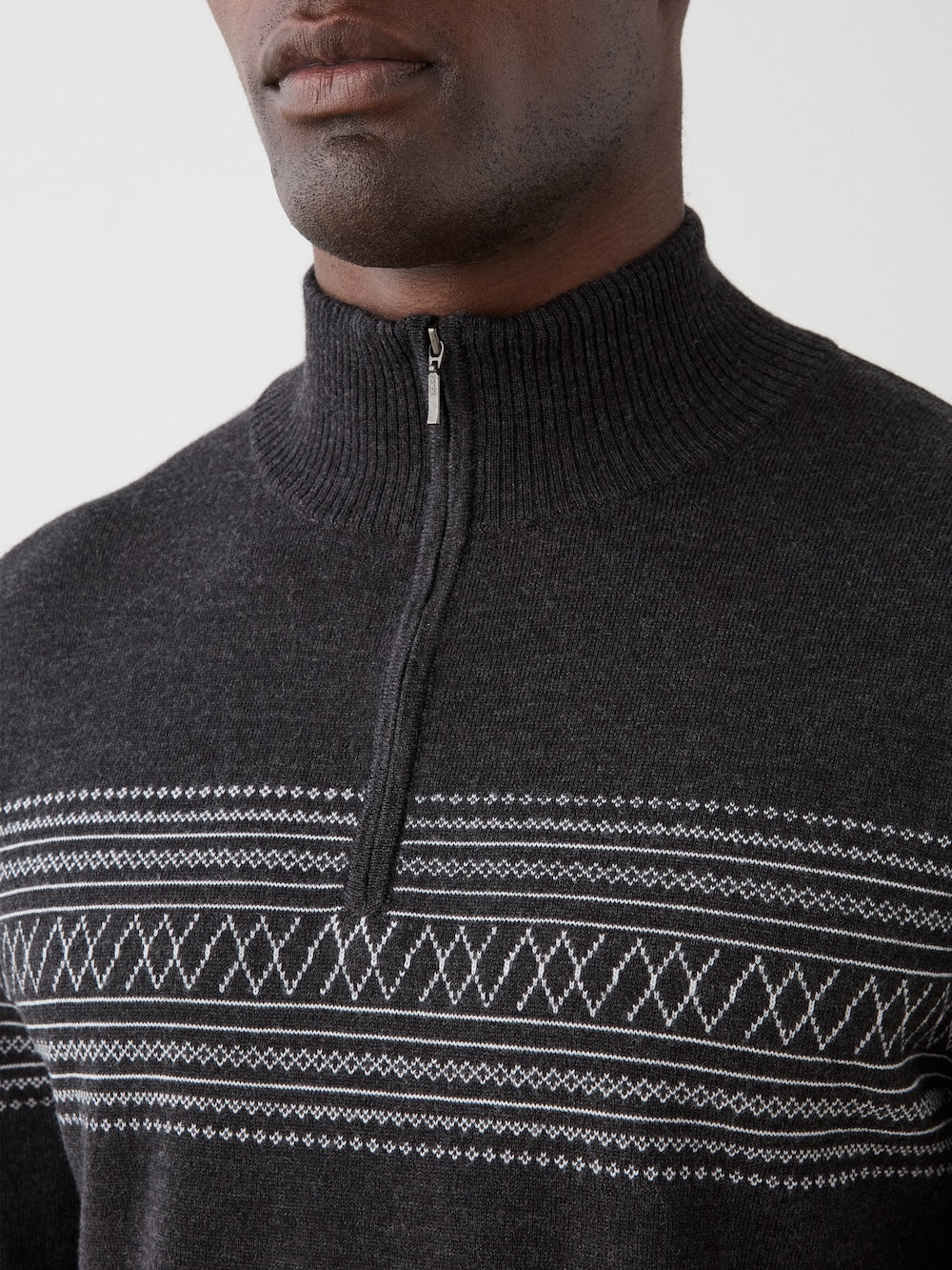 A person wearing the We Norwegians Signature Zip Up Men sweater, featuring a dark merino wool construction with a half-zip collar and a white Norwegian Setesdal pattern across the chest, shown from the shoulders up.