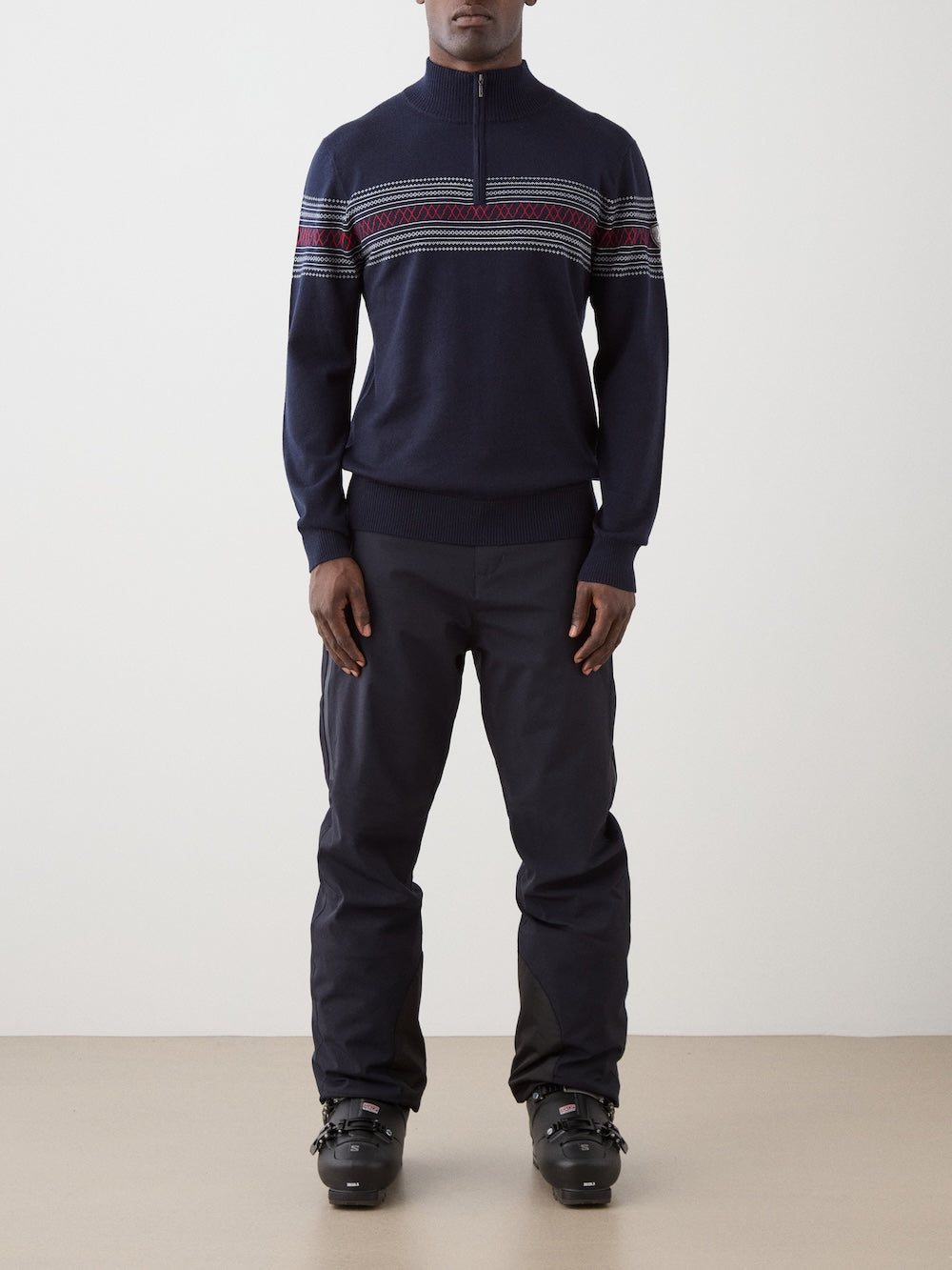 A person stands against a plain light background, wearing the We Norwegians Signature Zip Up Men—a navy blue sweater adorned with a classic red and white Norwegian Setesdal pattern, crafted from washable merino wool—paired with dark pants and black ski boots.