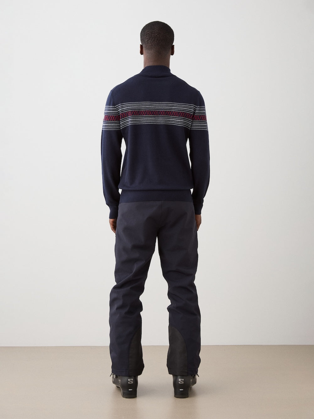 A person stands facing away, wearing the We Norwegians Signature Zip Up Men—a navy sweater featuring patterned stripes in the classic Norwegian Setesdal design across the upper back—paired with dark pants. The floor is beige and the background is plain white.
