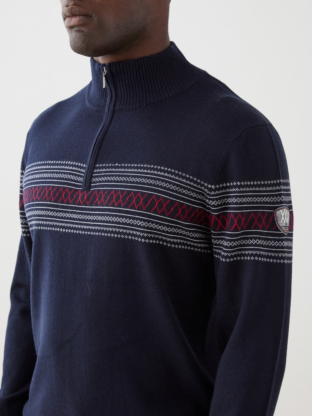 A person wearing the We Norwegians Signature Zip Up Men, a navy blue washable merino wool sweater decorated with red and white Norwegian Setesdal patterns across the chest and sleeves, stands against a plain light background.