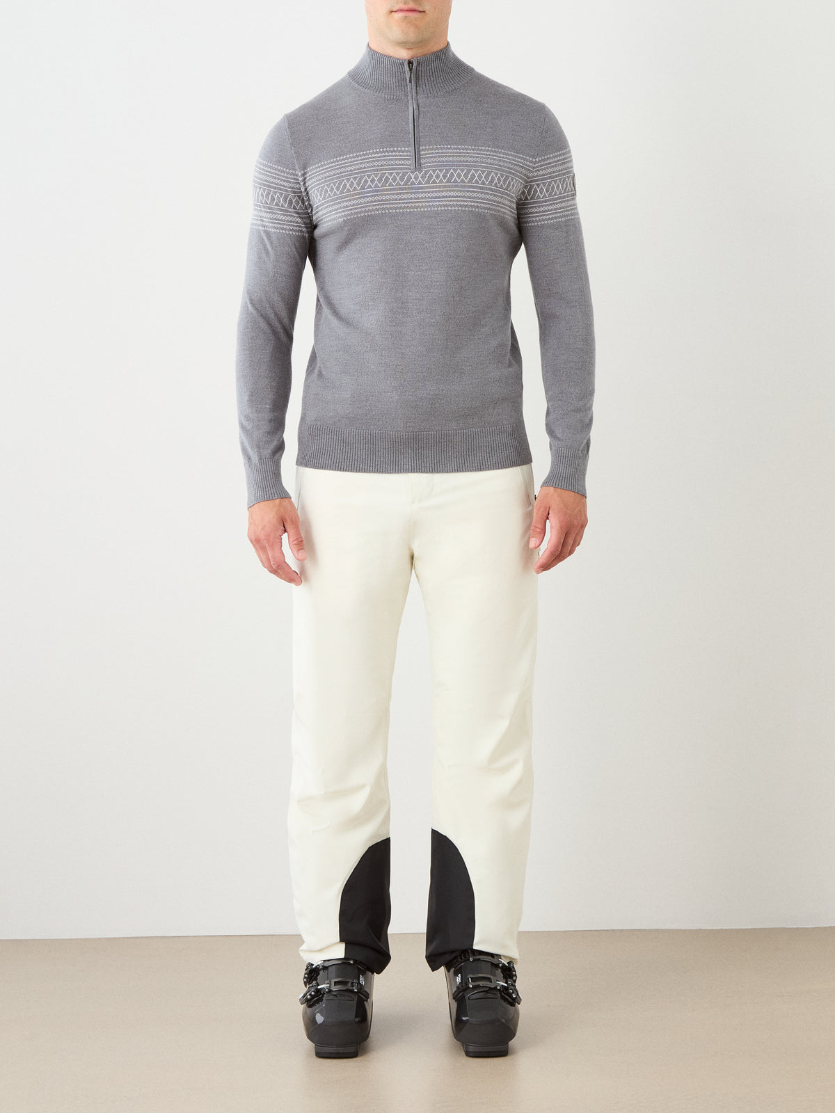 A person wearing the We Norwegians Signature Zip Up Men, a gray merino wool sweater featuring a subtle Norwegian Setesdal pattern, along with white pants with black patches at the ankles and black ski boots, stands against a plain light background.