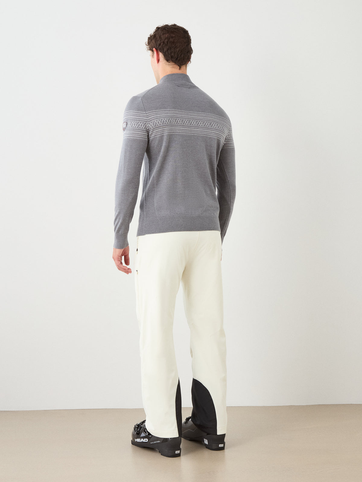 A person stands facing away, wearing the We Norwegians Signature Zip Up Men— a gray washable merino wool sweater featuring a Norwegian Setesdal pattern band across the back—paired with white pants and black shoes with white soles. The background is plain and neutral.