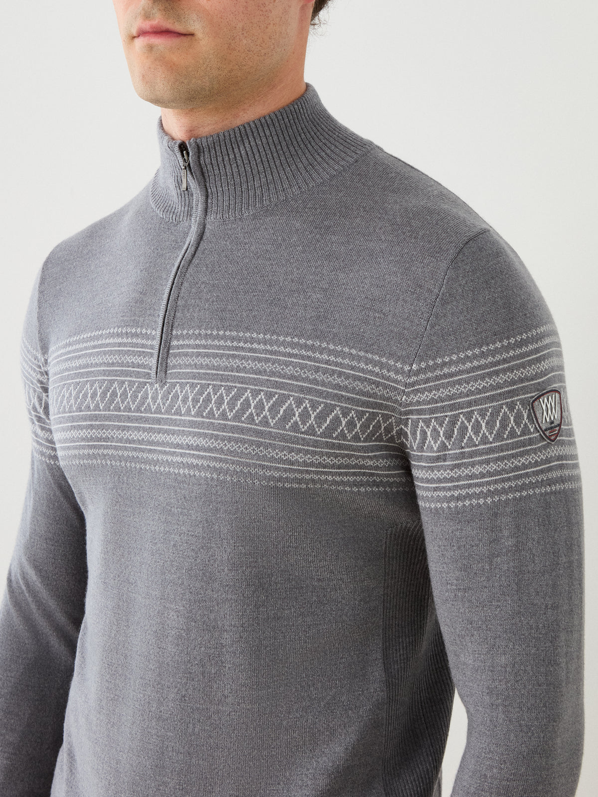 A person wearing the We Norwegians Signature Zip Up Men sweater—a light gray, long-sleeve merino wool zip-up with a half-zip collar and a white geometric Norwegian Setesdal pattern across the chest and upper arms. A small logo patch is visible on the left sleeve.