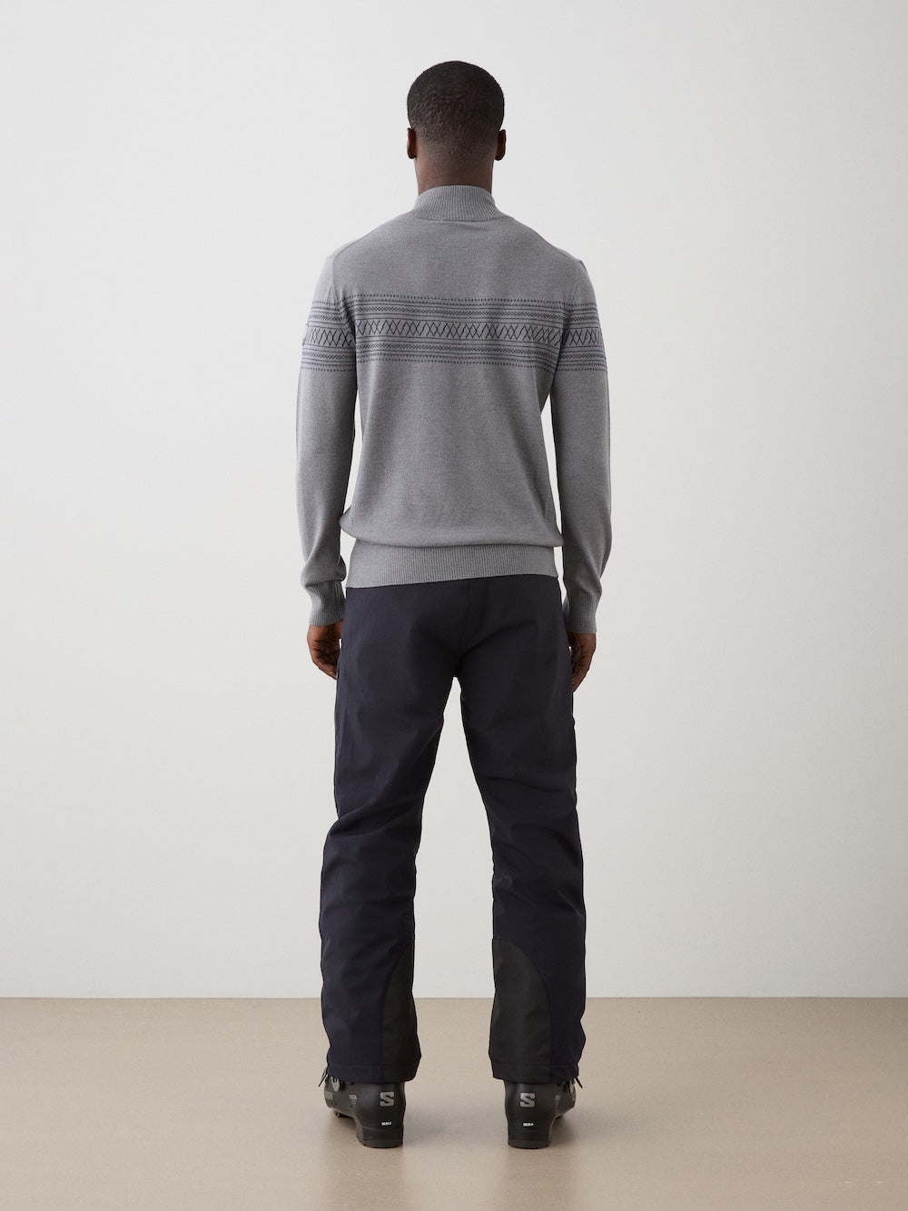 Person wearing The We Norwegians Signature Zip Up Men is a grey Merino wool sweater with a quarter-zip collar, dark Norwegian Setesdal patterns on the chest and arms, ribbed cuffs and hem, and a subtle logo patch on the left sleeve.