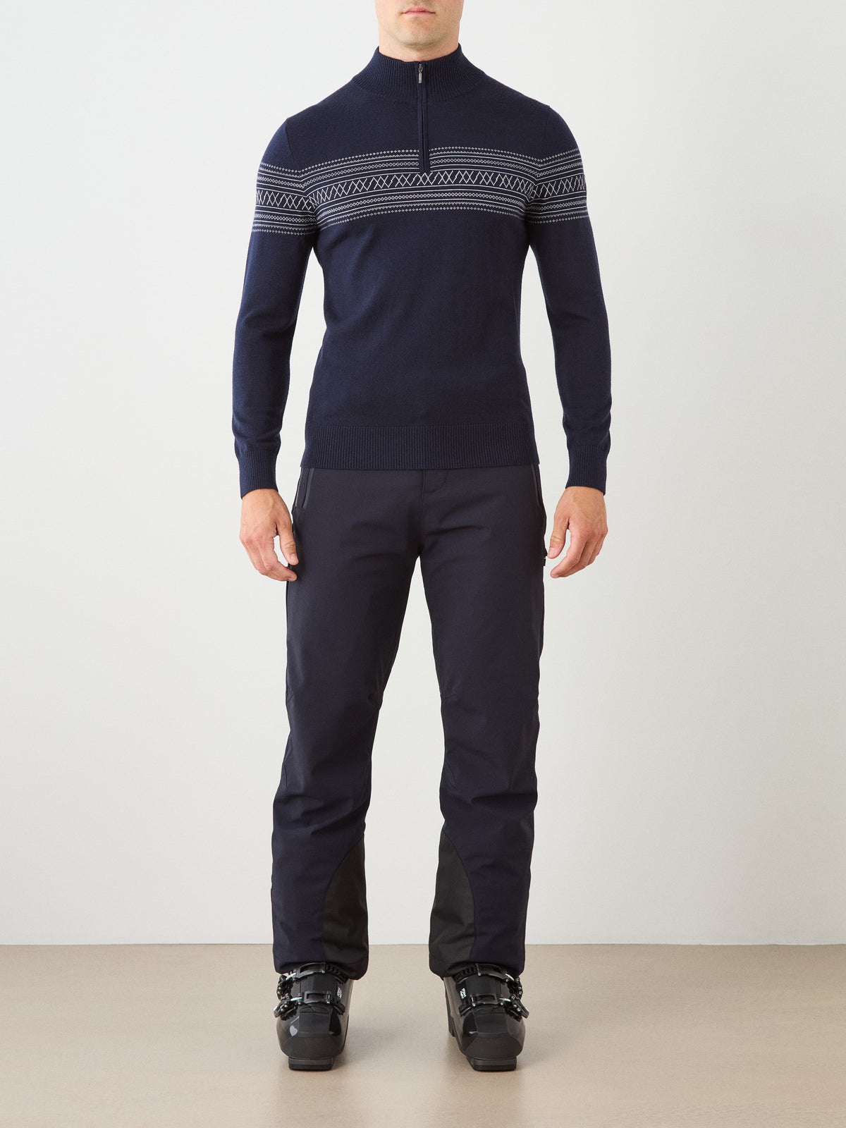 A person is wearing the We Norwegians Signature Zip Up Men ski sweater in navy blue merino wool with a white Norwegian Setesdal pattern, paired with dark ski pants and black ski boots, standing facing forward against a plain background.