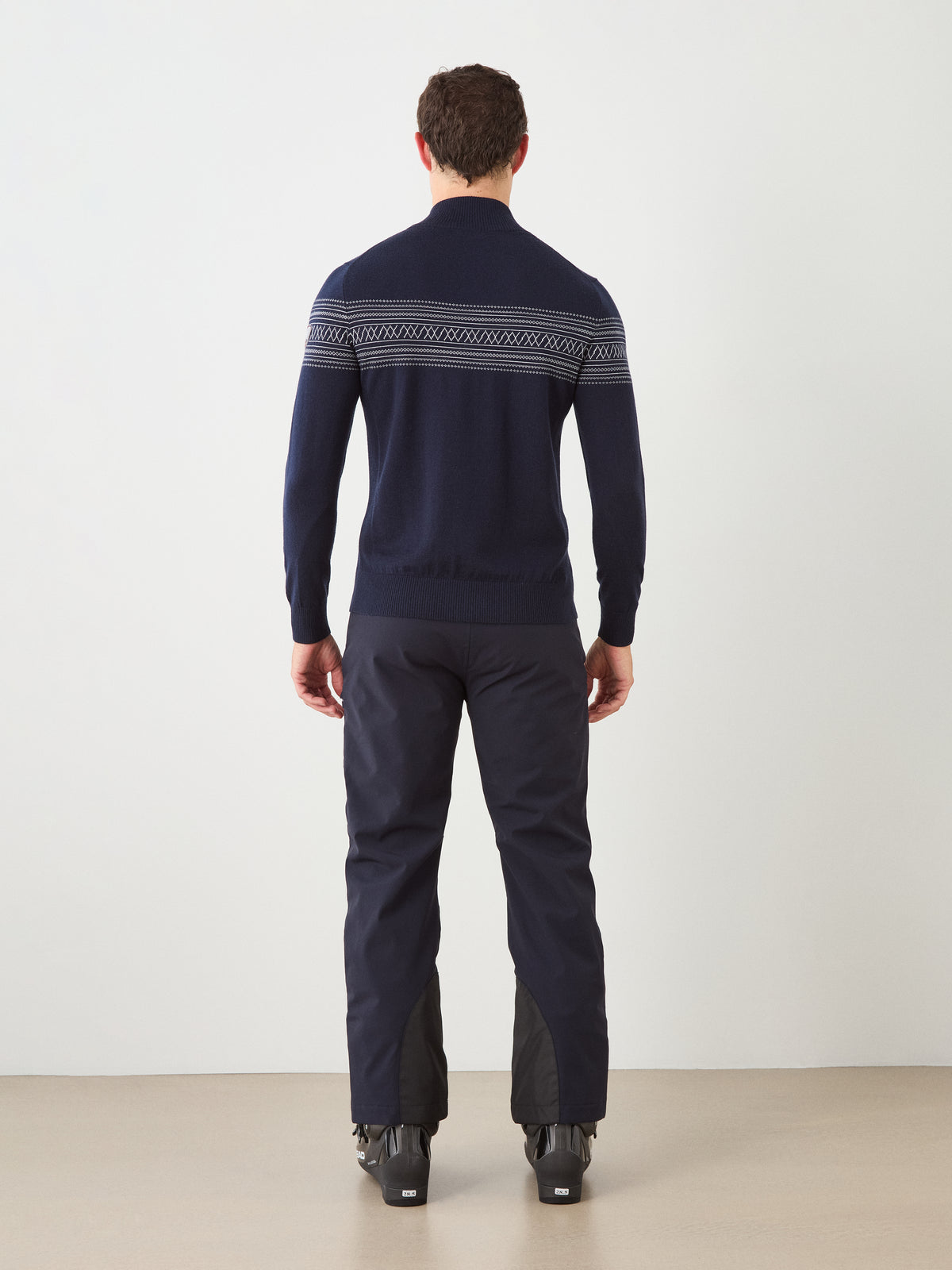 A person stands indoors facing away, wearing the We Norwegians Signature Zip Up Men—a navy merino wool sweater with a Norwegian Setesdal pattern across the upper back—paired with dark pants and black boots.