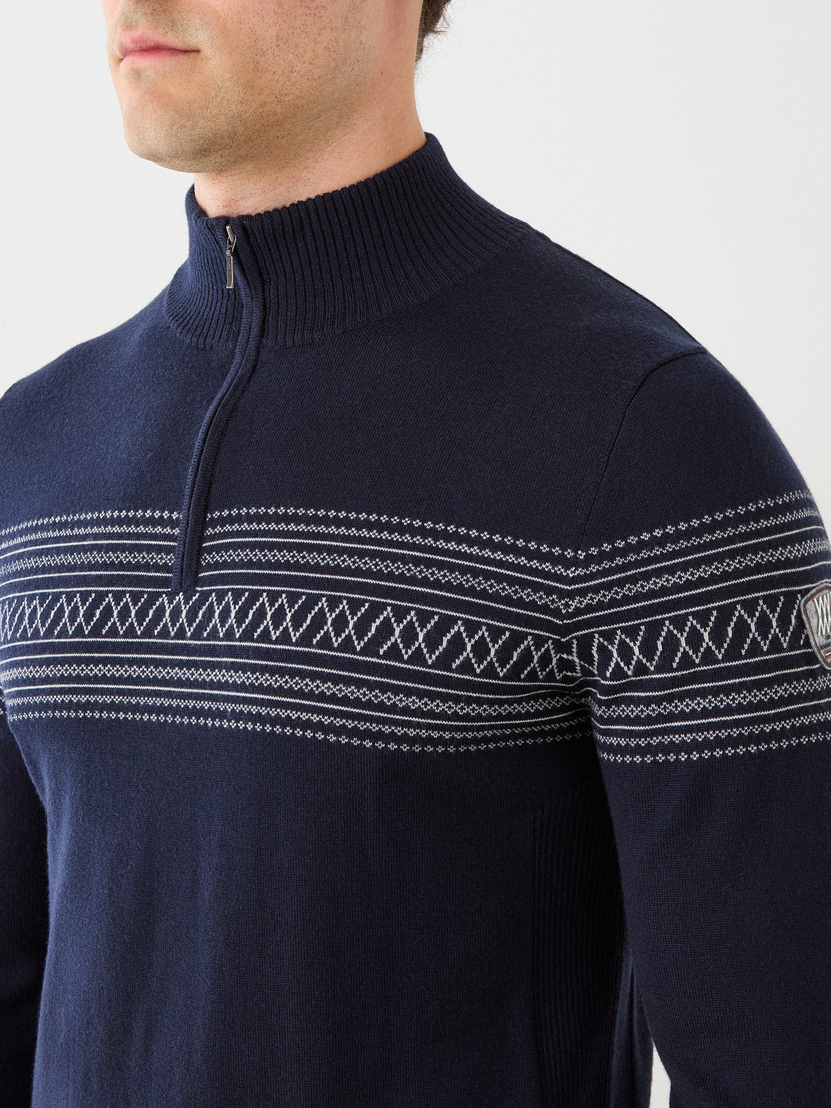 A man wearing We Norwegians’ Signature Zip Up Men sweater in navy blue merino wool, featuring white Norwegian Setesdal patterns across the chest and upper arms, stands against a plain light background.