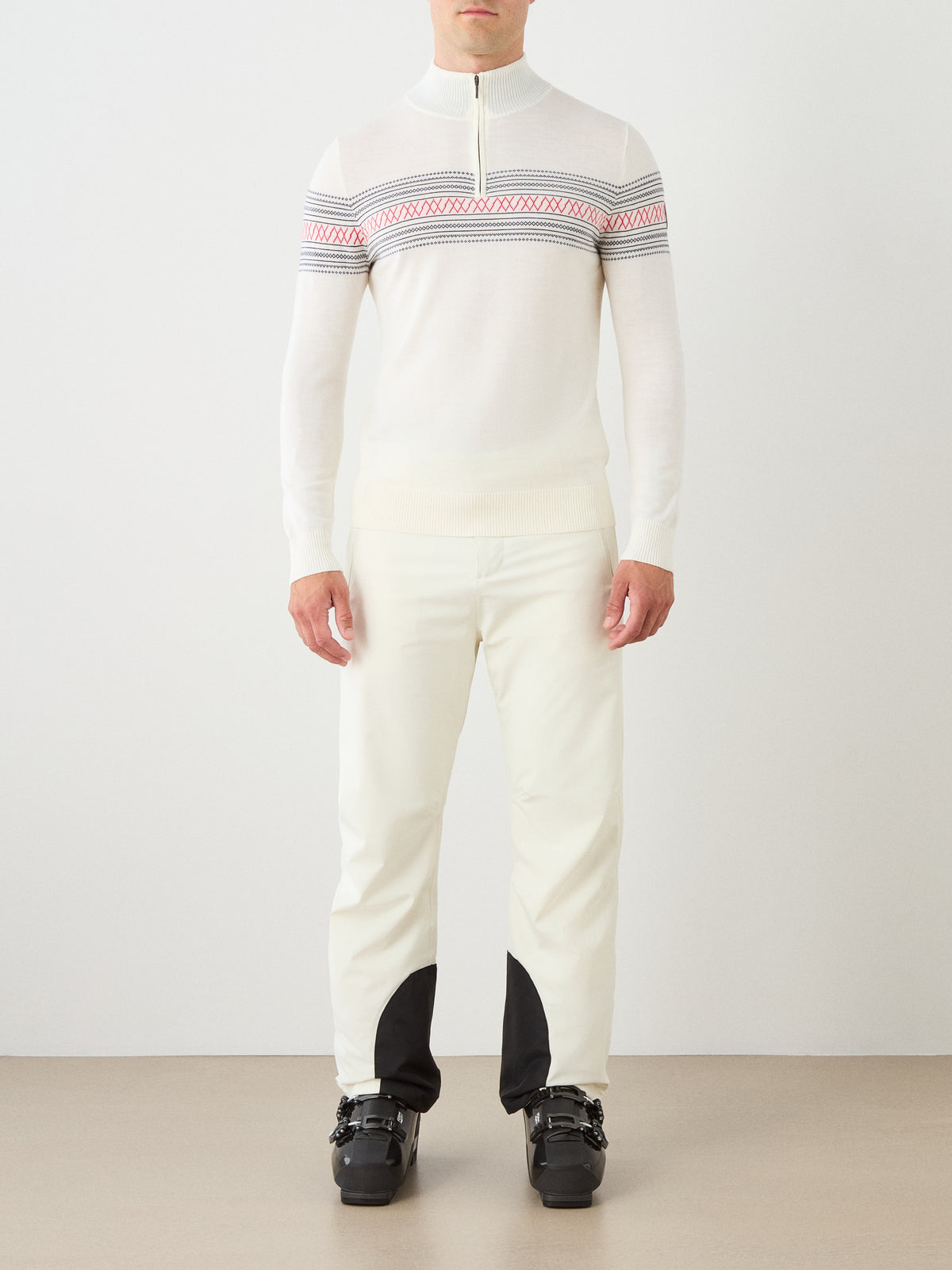A person wearing the We Norwegians Signature Zip Up Men—a white merino wool sweater featuring a red and gray Norwegian Setesdal pattern across the chest—paired with white pants that have black patches on the ankles and black ski boots, stands against a plain background.