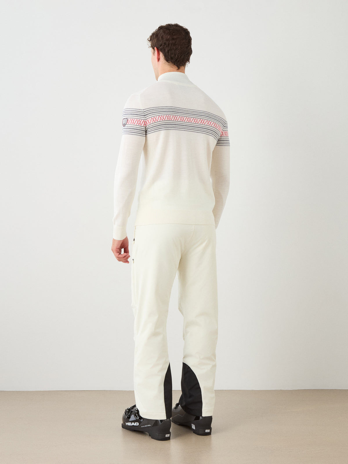 A man stands facing away in a minimalist indoor setting with a light background, wearing the We Norwegians Signature Zip Up Men—a white sweater featuring a Norwegian Setesdal pattern yoke—paired with white pants with black ankle accents and complemented by black ski boots.