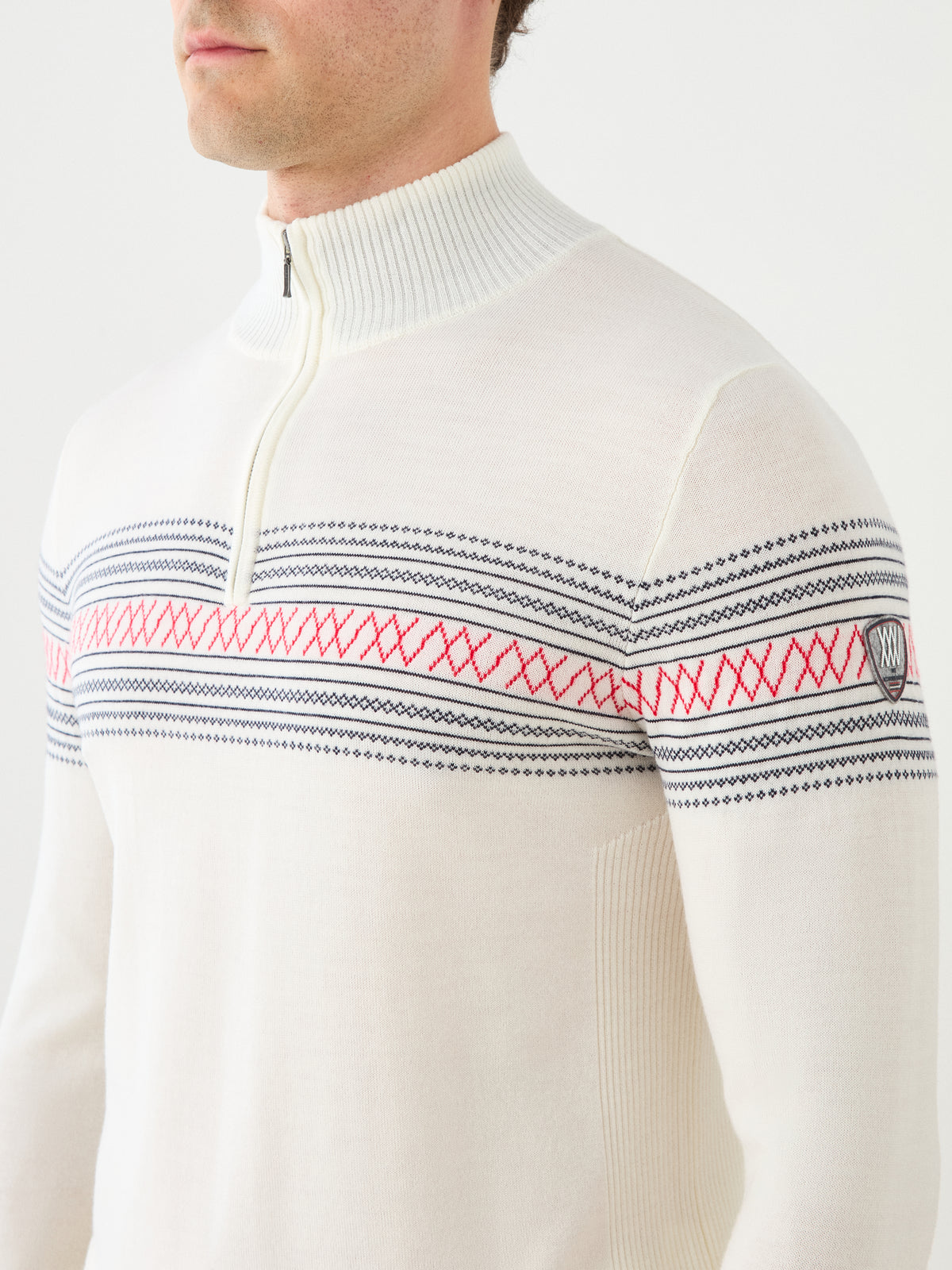A person wearing the We Norwegians Signature Zip Up Men—a white sweater featuring a half-zip collar and traditional Norwegian Setesdal-inspired black and red line and X pattern across the chest and upper arms, crafted from washable merino wool for comfort and easy care.