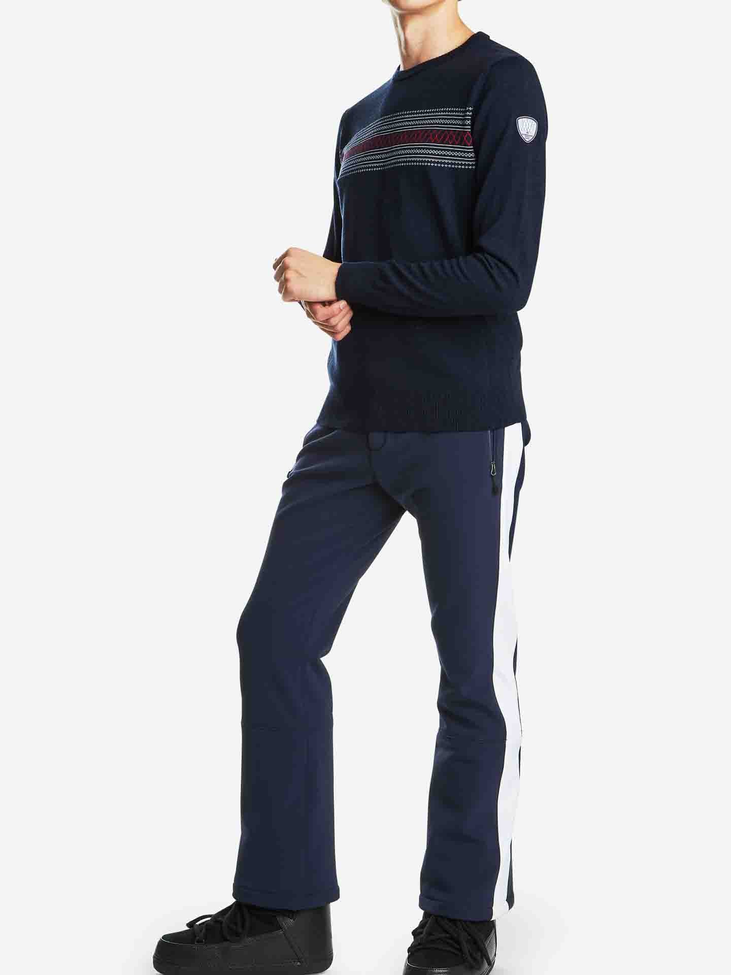 A person stands against a plain white background, wearing the Signature Crewneck Men by We Norwegians. The navy blue merino wool sweater features red, white, and blue stripes across the chest and a small patch on the left shoulder. They are also wearing navy blue pants with white side stripes and black shoes. Their arms are relaxed at their sides.