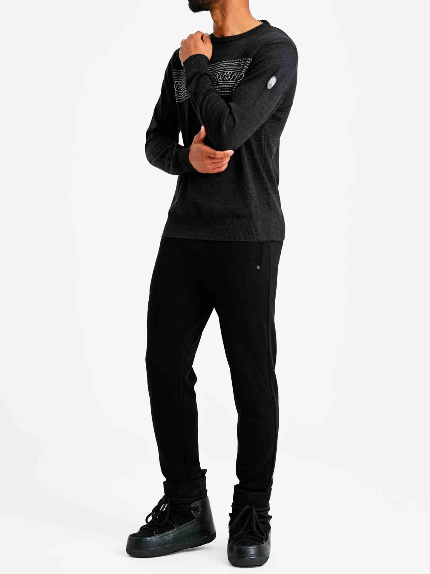 A person standing against a white background wearing the We Norwegians Signature Crewneck Men in dark gray merino wool, which features a geometric pattern reminiscent of Norwegian heritage. They are also dressed in black pants and black high-top sneakers. One hand is near their chin while the other rests by their side.