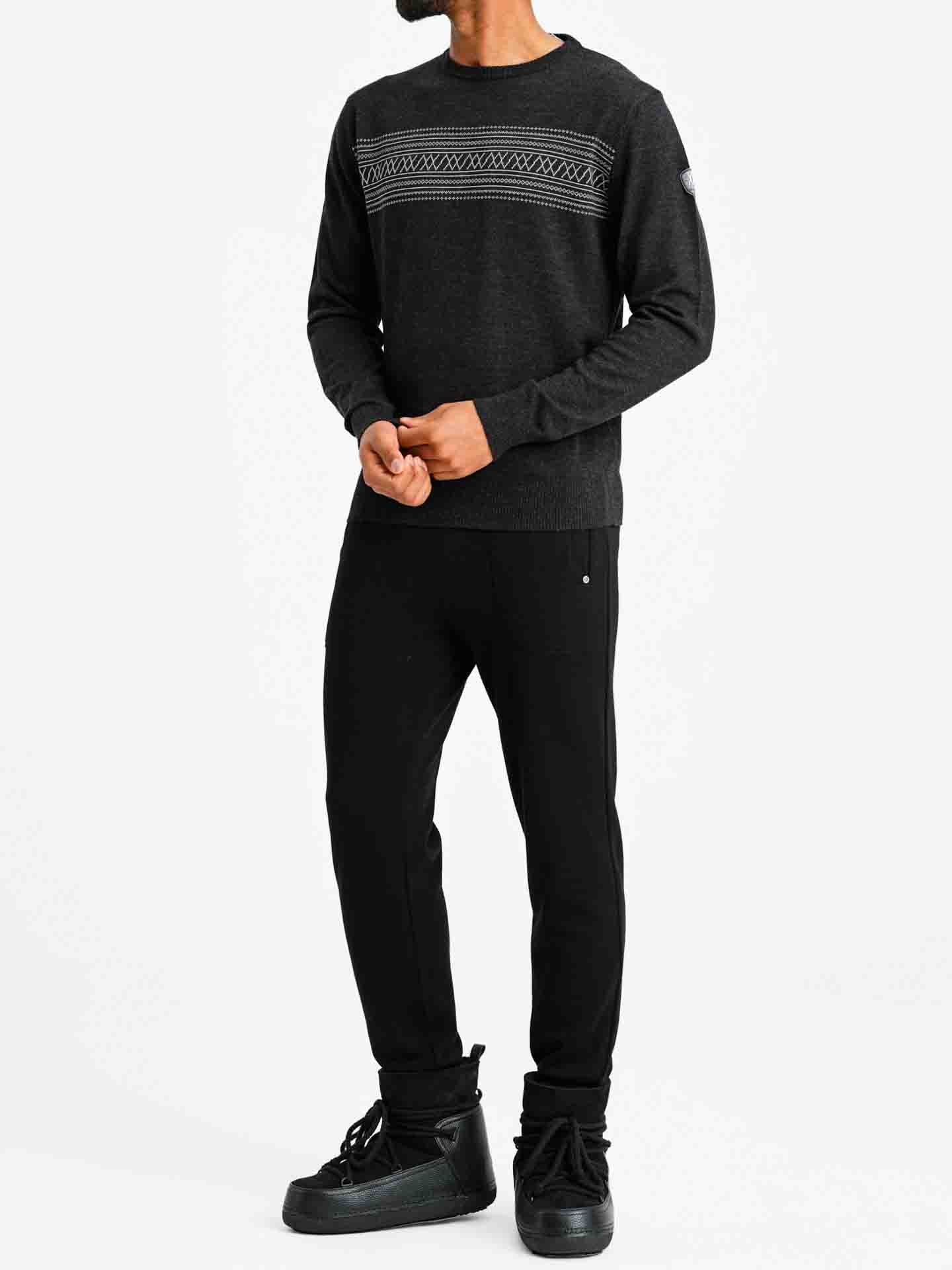 A person stands against a plain white background wearing the Signature Crewneck Men from We Norwegians, a dark, long-sleeved merino wool sweater featuring intricate patterns inspired by Norwegian heritage across the chest. They are also dressed in black pants and black high-top shoes, with their hands clasped in front of them.