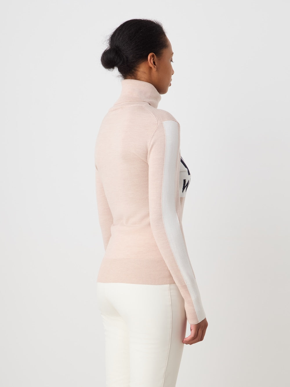 A woman with her hair in a bun wears the We Norwegians World Cup Sweater women - Pre Spring 26 in light pink with white side stripes and white pants, standing sideways and facing left against a plain white background.