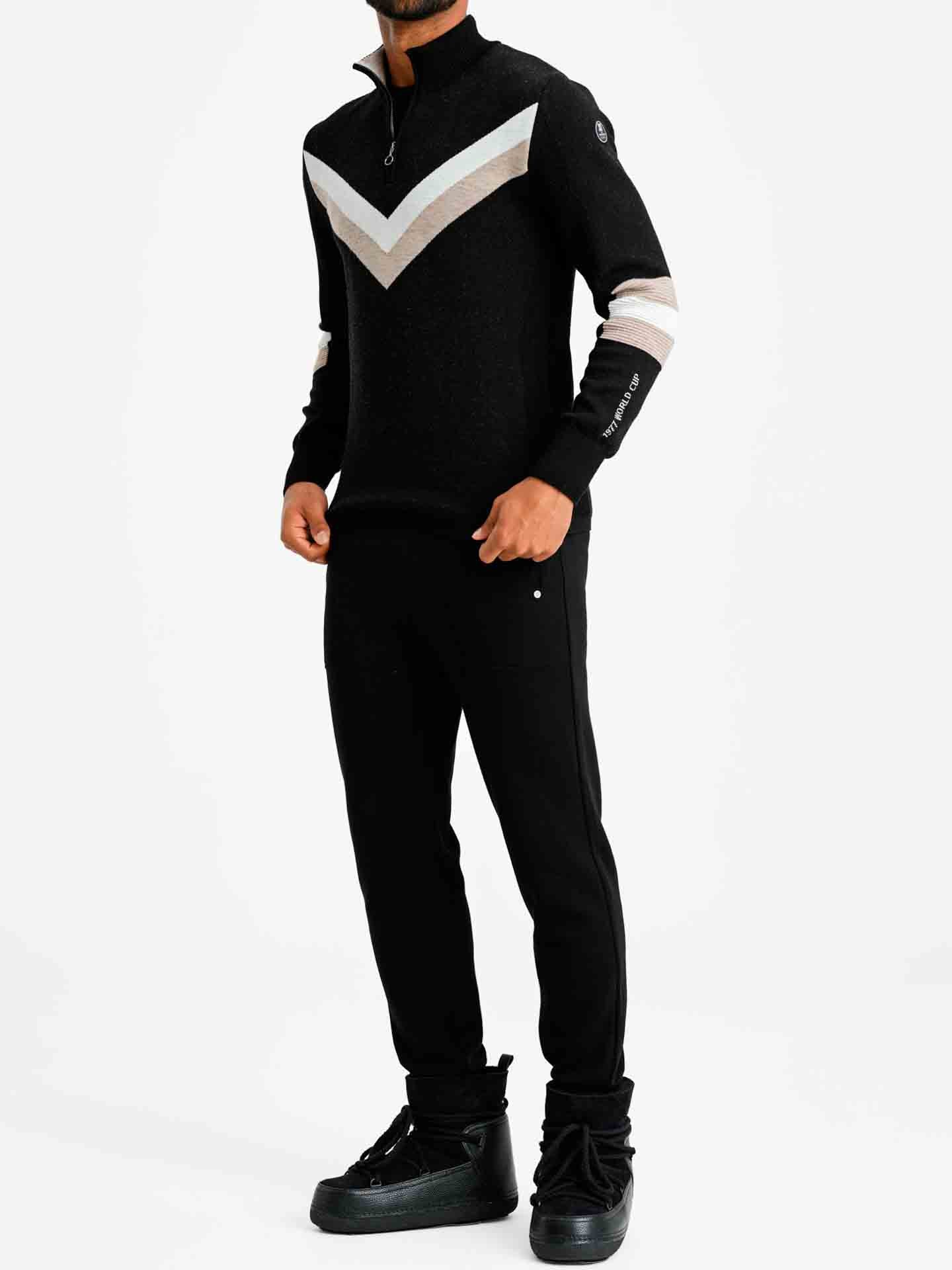 A person is standing in the Hafjell Zip Up Men by We Norwegians, a piece of black athletic wear made from 100% fine merino wool. The garment features a white and grey chevron pattern on the chest and upper arms, as well as a small zipper at the neck. They are wearing black sneakers and posing against a plain white background, with their head not visible in the frame.