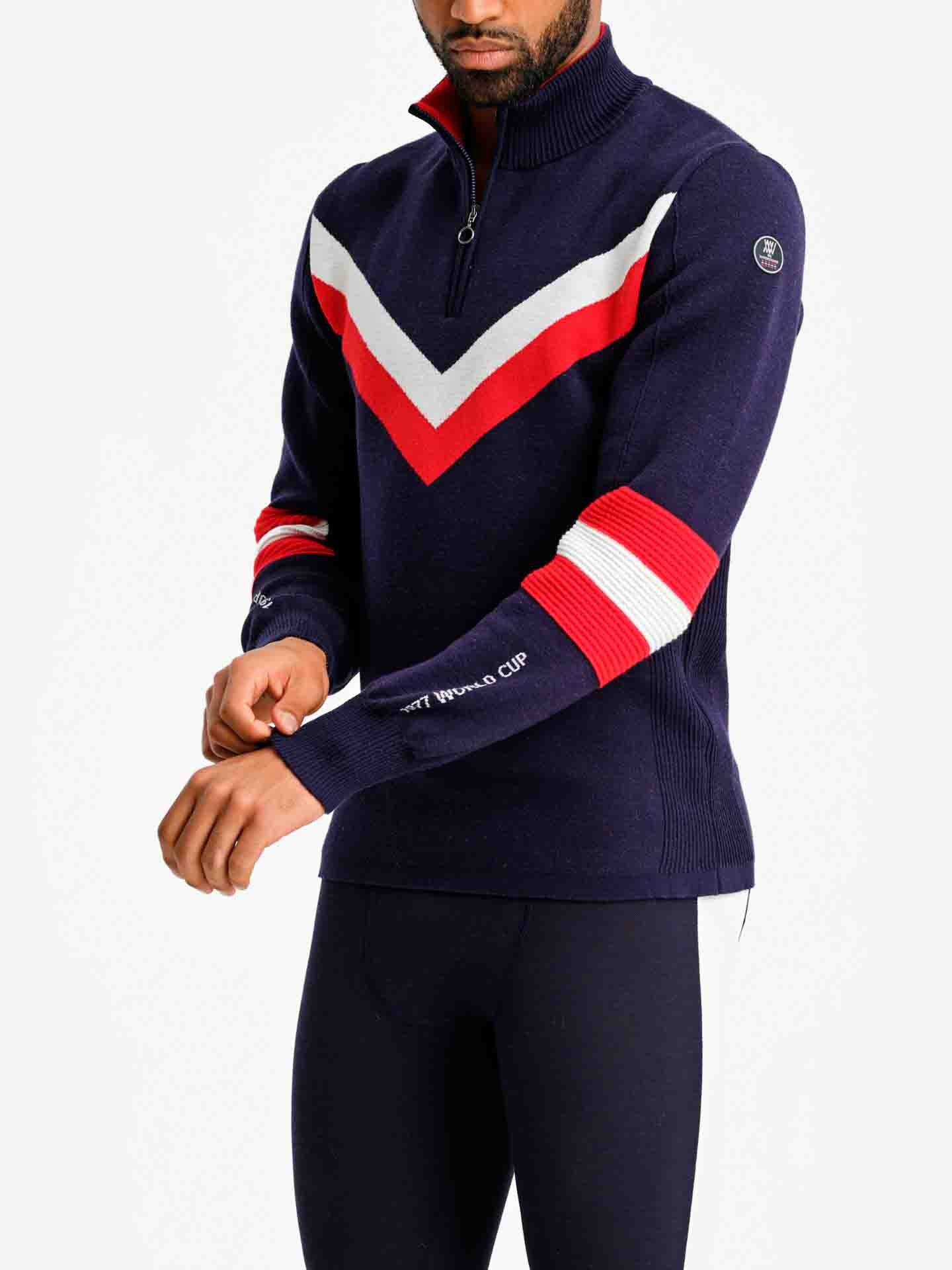 A person wearing the Hafjell Zip Up Men from We Norwegians, a navy blue ski sweater featuring red and white chevron stripes on the chest and multi-colored stripes on the sleeves. They are adjusting the cuff of one sleeve, which displays the text "34th PRADA CUP." This exquisite piece is crafted from 100% fine merino wool by Italian knitters. The pants are black.