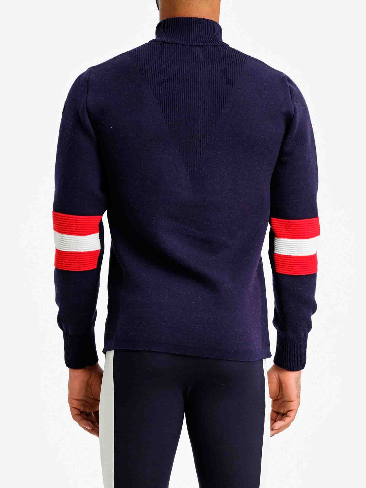 A person wearing the Hafjell Zip Up Men sweater, designed by We Norwegians, in dark blue with red and white stripes on the upper arms, crafted from 100% fine merino wool by Italian knitters, along with navy pants, is shown from the back. The ski sweater features a ribbed collar and a V-shaped ribbed design on the back, set against a plain white background.