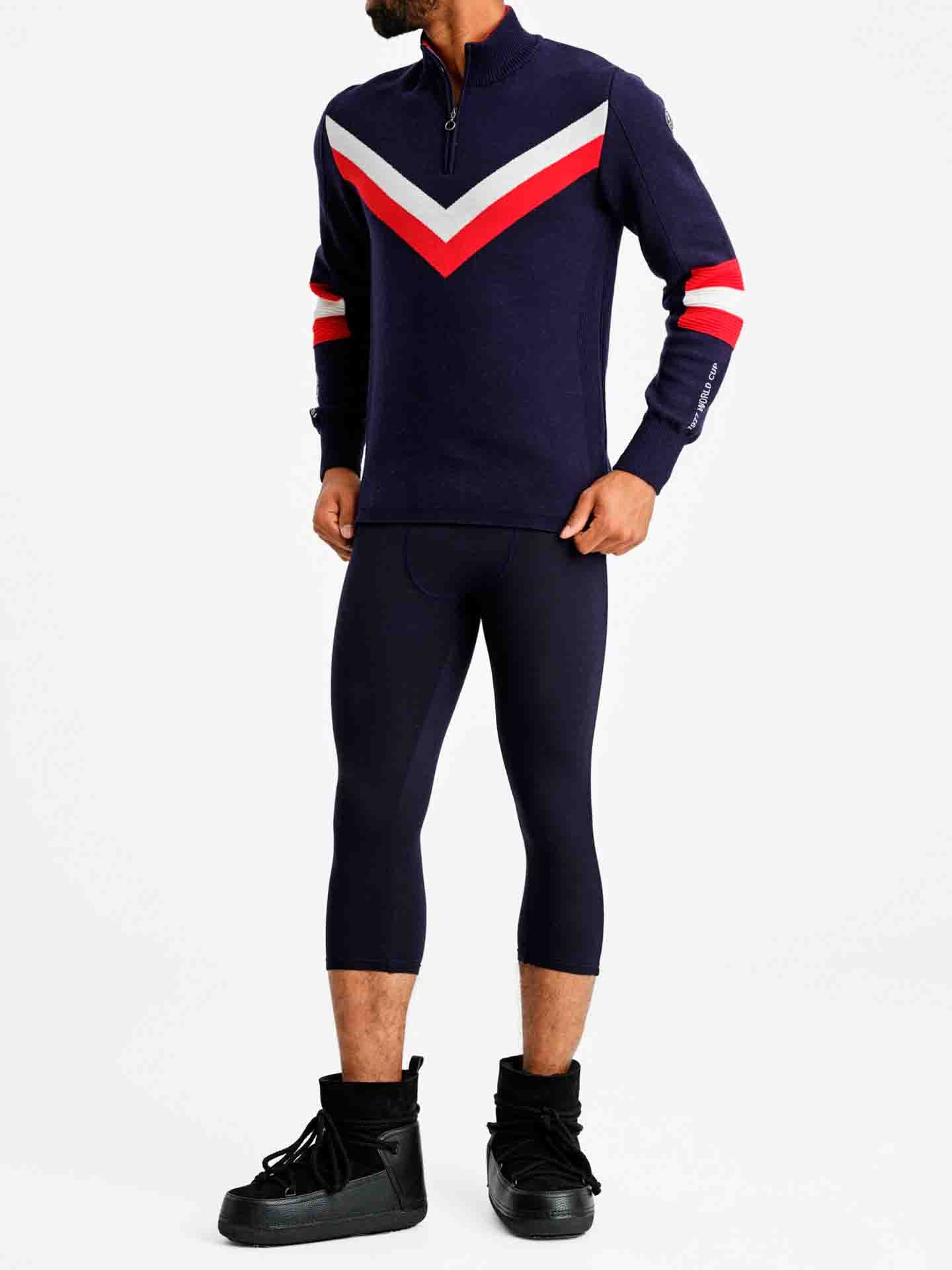 Person wearing the Hafjell Zip Up Men by We Norwegians, a long-sleeve navy blue top with red and white chevron stripes, reminiscent of classic ski sweaters made by Italian knitters, paired with black three-quarter length leggings and black high-top sneakers. The individual stands against a plain white background.