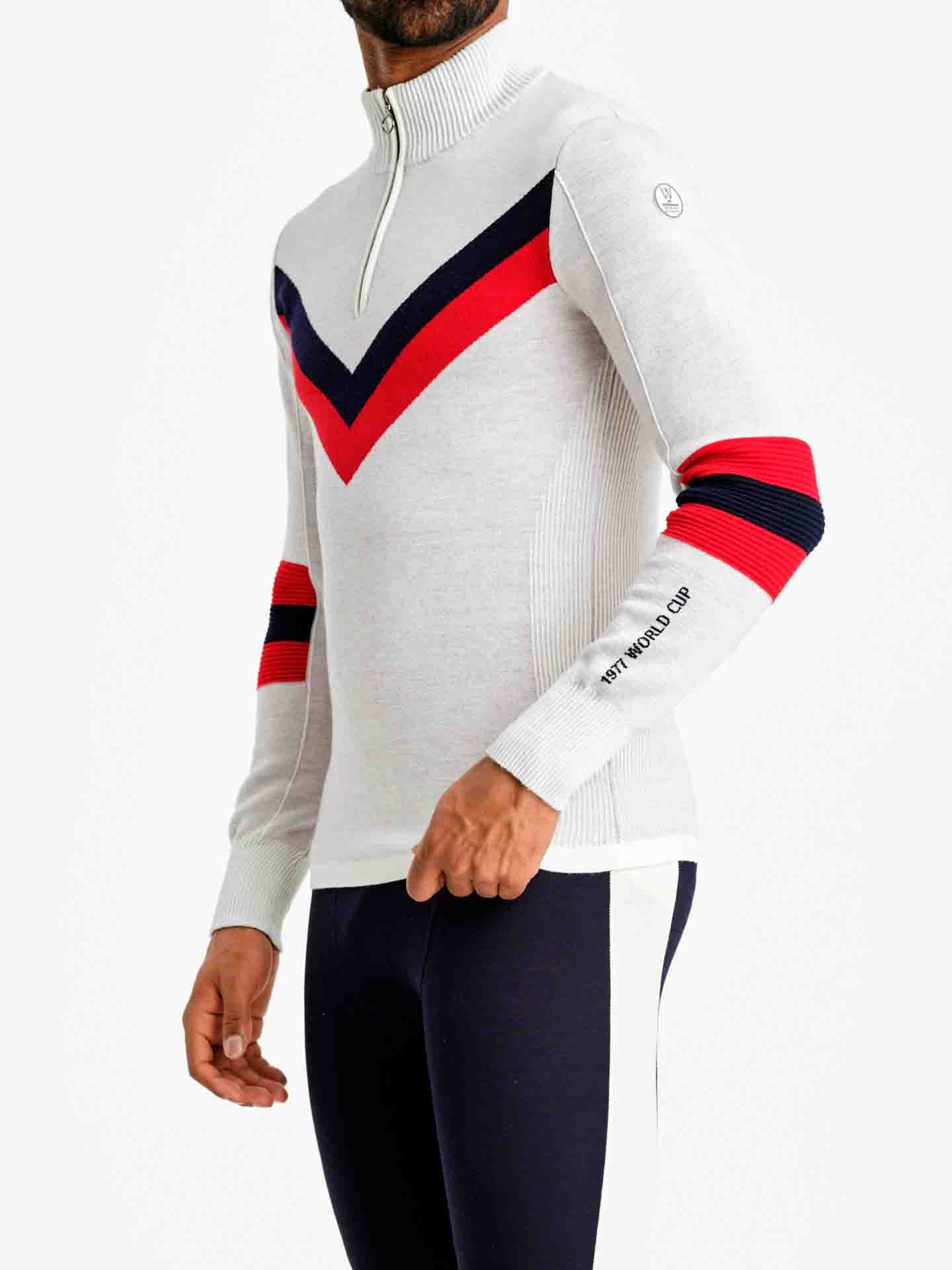 A person models the Hafjell Zip Up Men by We Norwegians, a fitted long-sleeve athletic top with a high zipper collar, reminiscent of stylish ski sweaters. The top is light gray with red and black chevron stripes across the chest and similar striped details on the sleeves, crafted beautifully by Italian knitters. The person pairs it with black sporty pants featuring white side stripes.