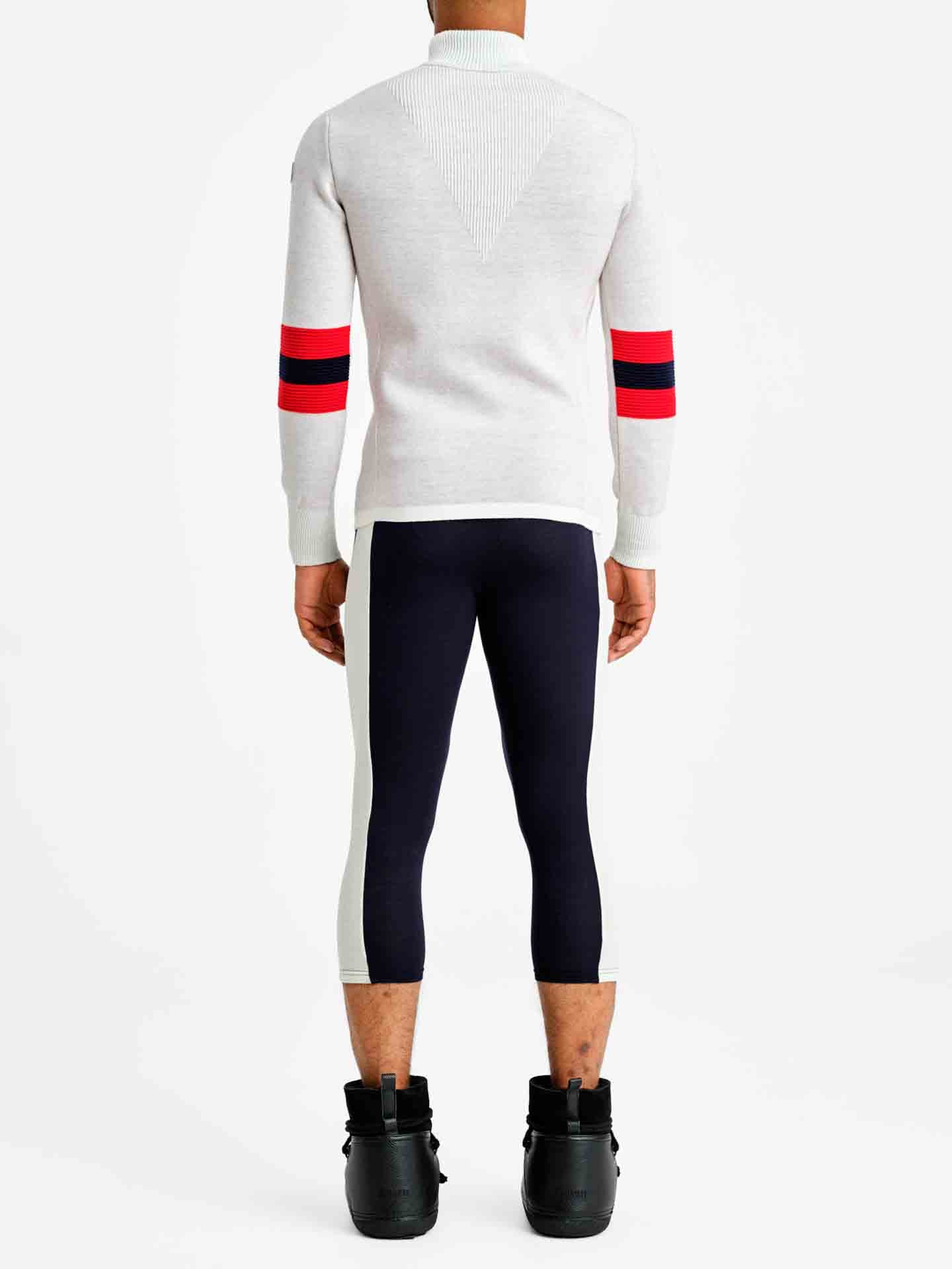 A person wearing the Hafjell Zip Up Men, a white sweater with red and blue stripes on the sleeves crafted by Italian knitters using 100% fine merino wool from We Norwegians, along with black and white leggings and black high-top sneakers, stands facing away from the camera against a plain white background.