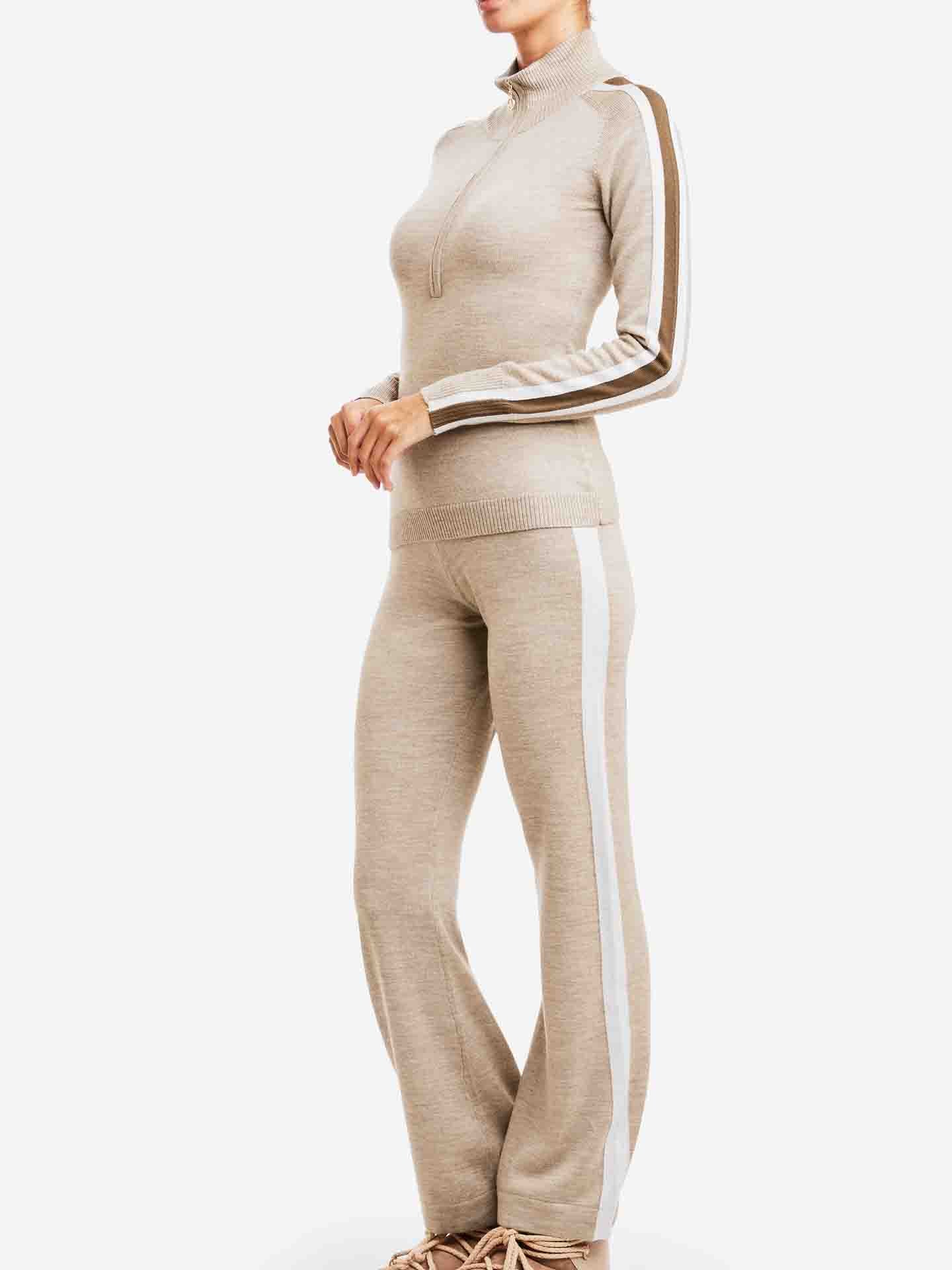 A person wearing a Merino Wool beige knit tracksuit from We Norwegians, featuring white and brown stripes down the sleeves and sides of the pants, stands against a plain white background. The aprés ski outfit includes a long-sleeve, high-neck Geilo Zip Up Women top and matching pants.