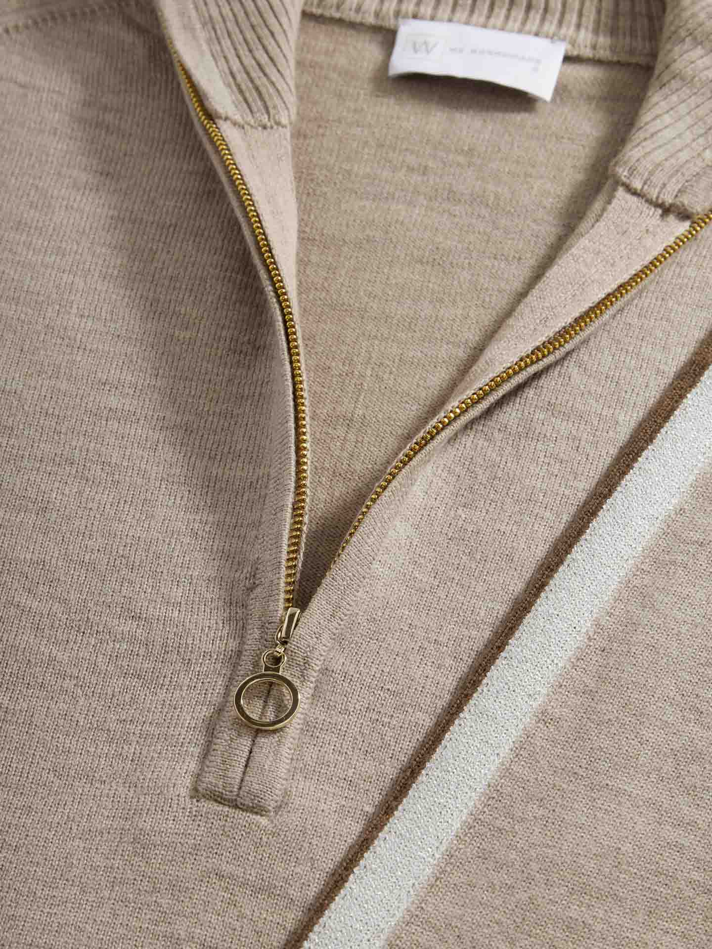 Close-up of a beige Geilo Zip-Up Women sweater by We Norwegians made of fine knit Merino Wool. The aprés ski-inspired sweater features a brass zipper partially undone, with a circular zipper pull. A white and light brown diagonal stripe is visible on the right side of the image.