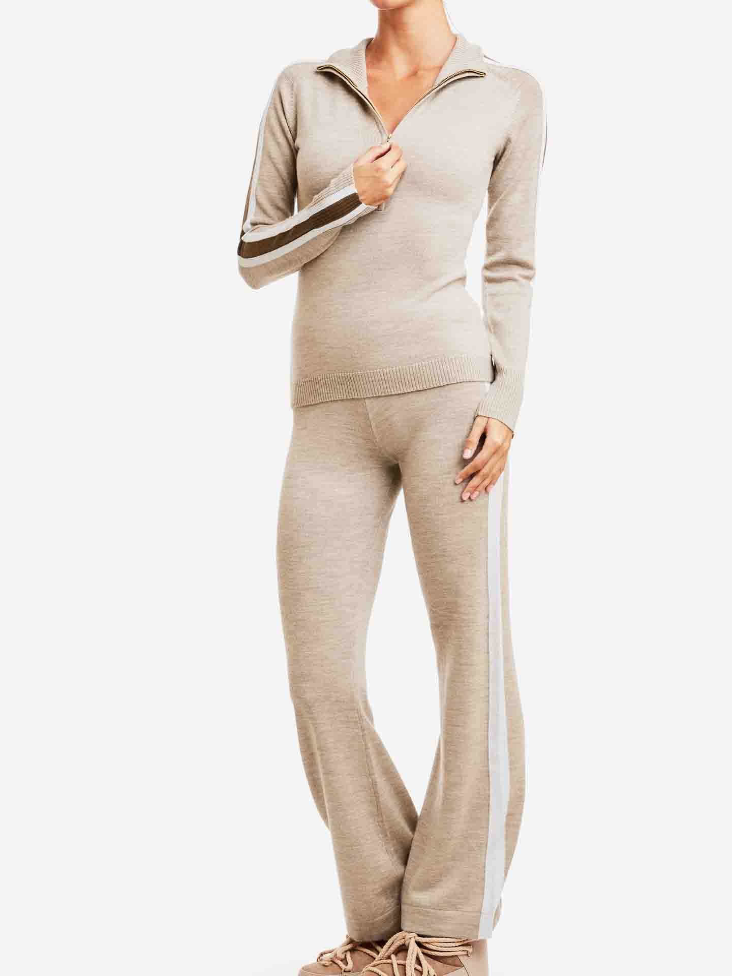 A person is posed against a white background, wearing a beige We Norwegians Geilo Zip Up Women tracksuit featuring white stripes on the arms and legs. The ensemble includes a long-sleeve zip-up top and matching pants. The person has one hand lightly holding the zipper of the top, and beige shoes are visible.