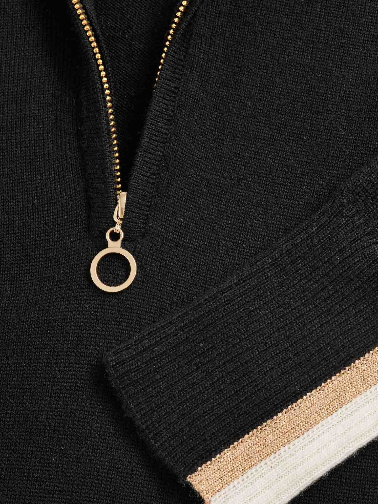 Close-up of the We Norwegians Geilo Zip-Up Women garment in black, showcasing a gold zipper with a circular pull and a sleeve featuring white and gold striped cuffs. Crafted from soft Merino wool, the visible knit texture makes it ideal for an après-ski look.