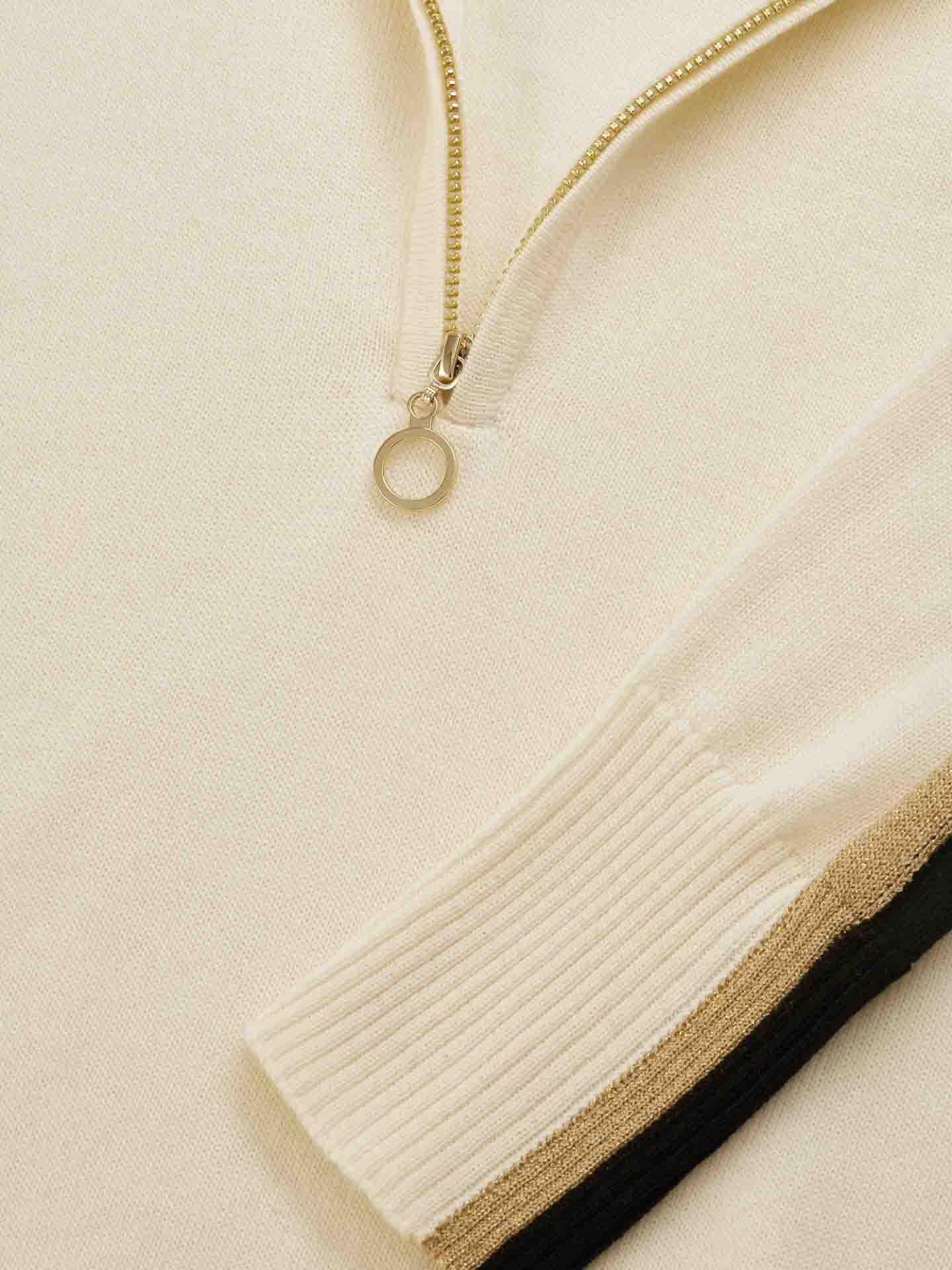 Close-up of a beige Geilo Zip Up Women garment from We Norwegians with a zipper partially undone. The zipper features a circular pull tab, and the sleeve cuff is ribbed. A narrow black and brown stripe accents the sleeve. The soft fabric appears to be knit, possibly Merino Wool, making it perfect for après ski lounging.