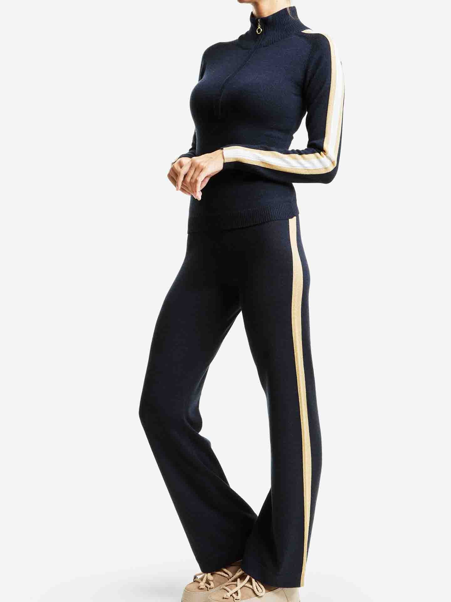 A standing individual wearing the We Norwegians Geilo Zip Up Women tracksuit, featuring a black zip-up top and straight-leg pants with white and gold stripes along the sides of both the sleeves and pants. They are also wearing beige athletic shoes, evoking après ski vibes. The background is plain white.