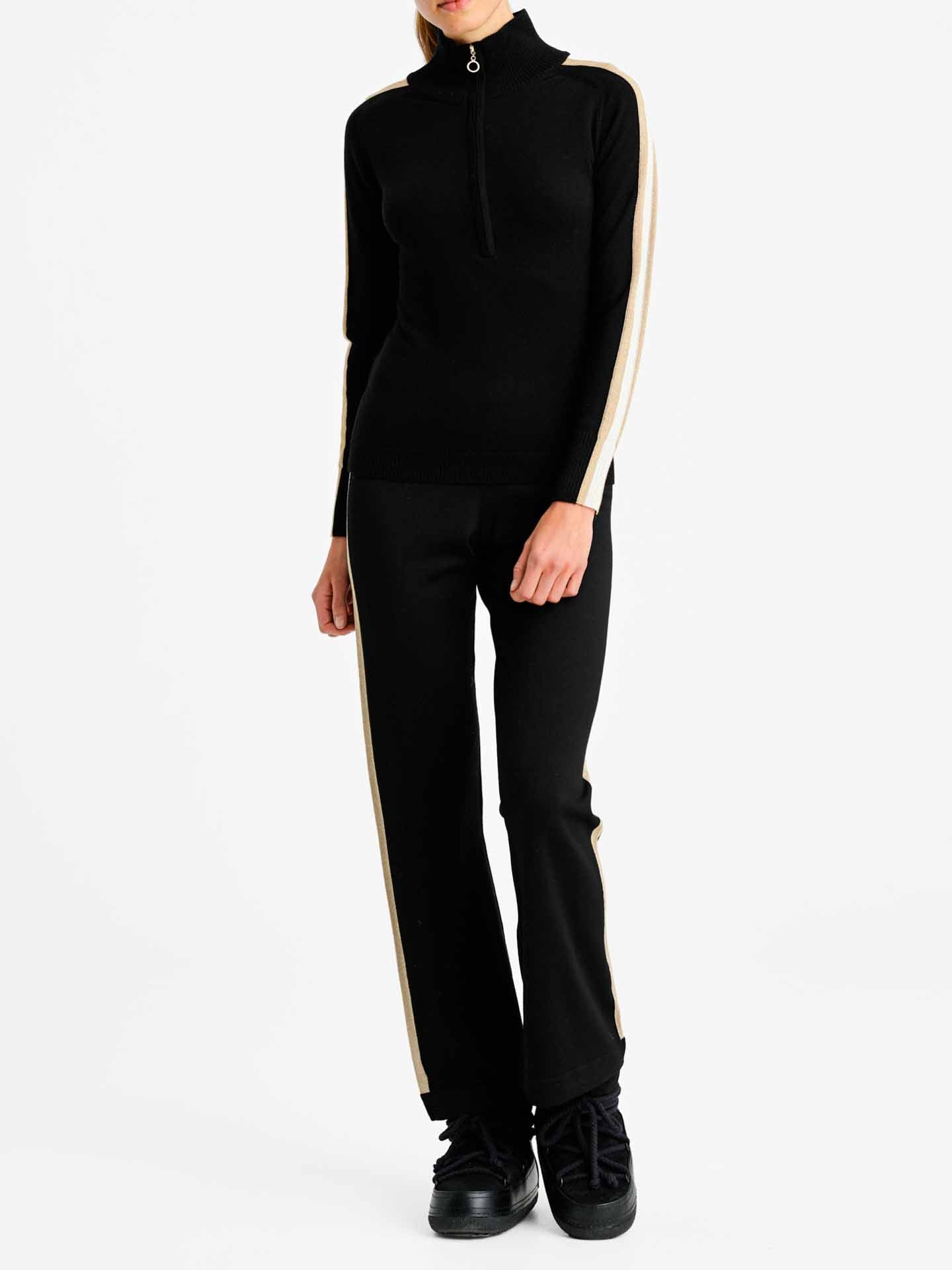 A person is standing against a white background wearing the Geilo Zip Up Women tracksuit by We Norwegians, featuring a quarter-zip top and matching pants in black with beige stripes on the sleeves and sides. The ensemble, reminiscent of an apres ski look, is completed with black sneakers.