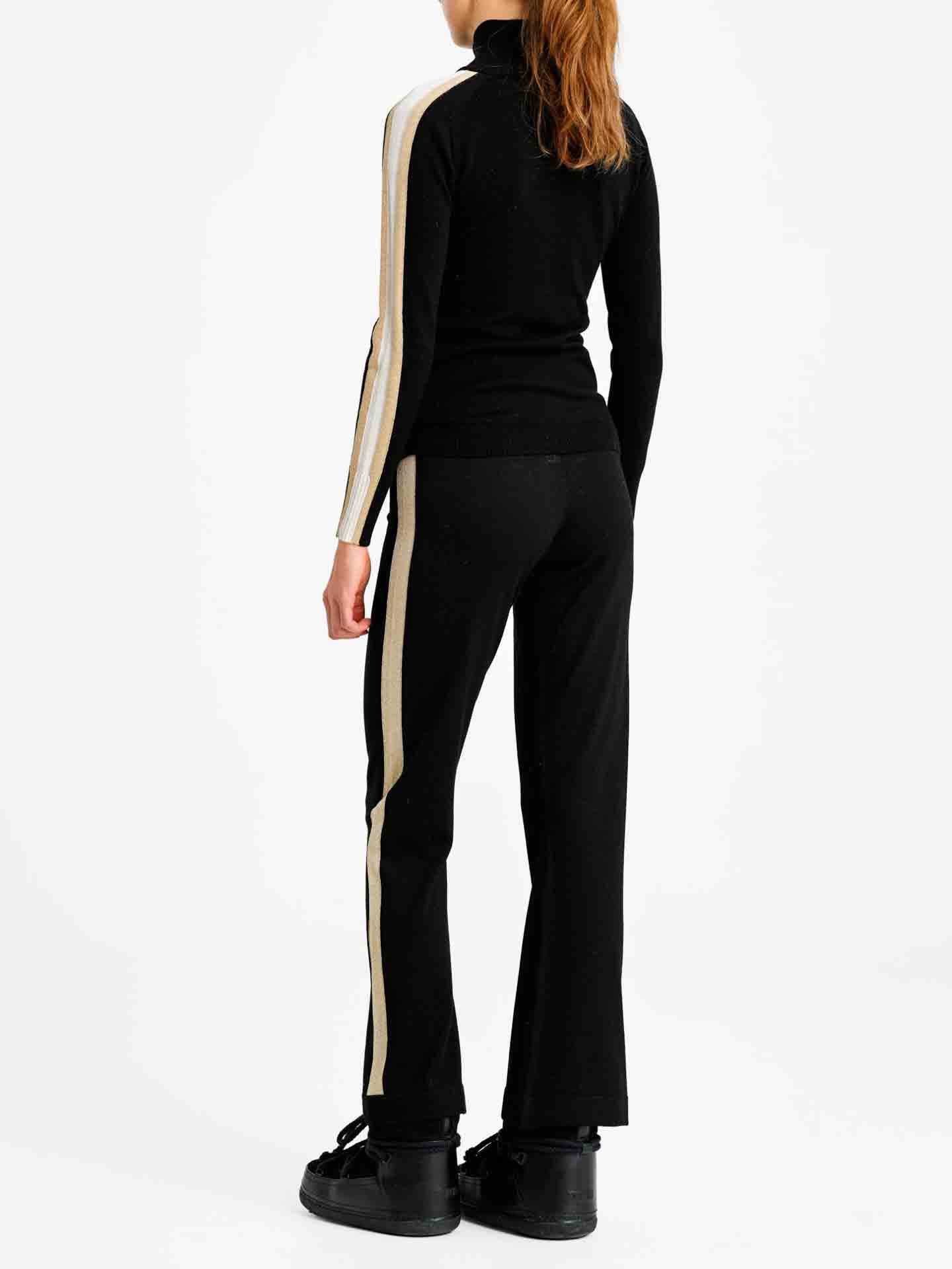 A person with long hair stands facing away, wearing the Geilo Zip Up Women by We Norwegians, a black jacket with beige stripes down the sleeves and matching pants. The outfit, perfect for an après-ski look, is paired with black shoes. The background is white and plain.