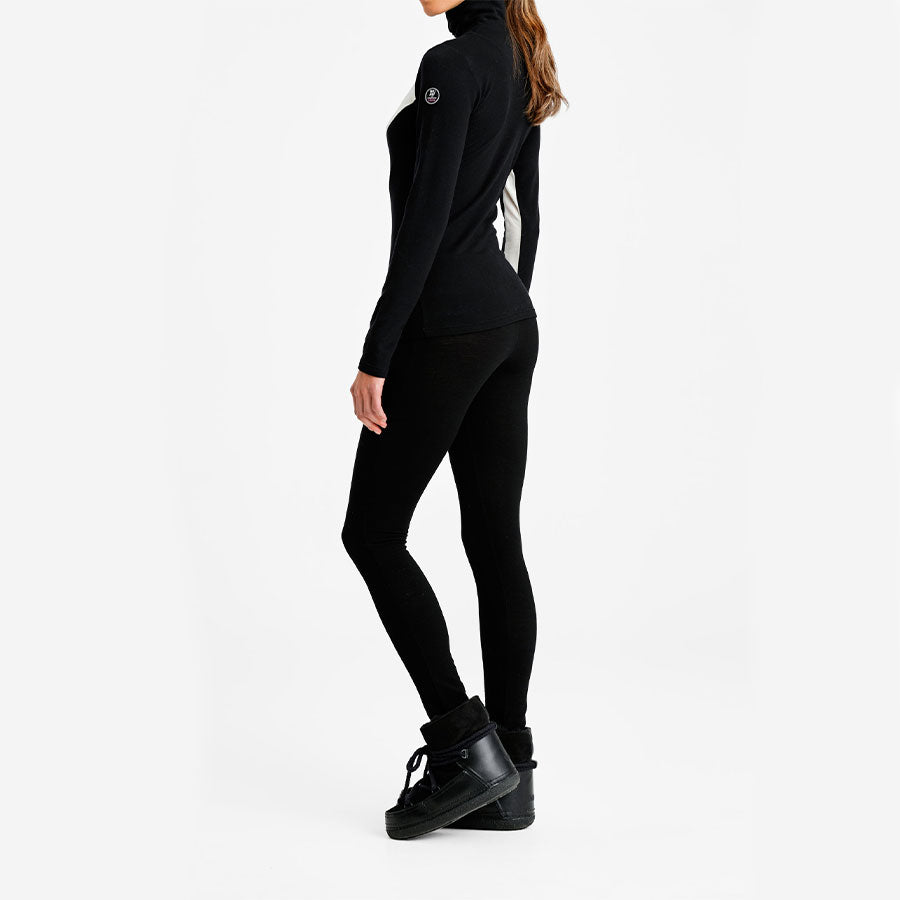 A woman is standing sideways, exuding retro vibes in the We Norwegians Voss Chevron ZipUp Women, a black fitted turtleneck sweater made from Super Fine Merino wool with a visible logo on the sleeve, paired with black leggings and black ankle boots. The background is plain white.