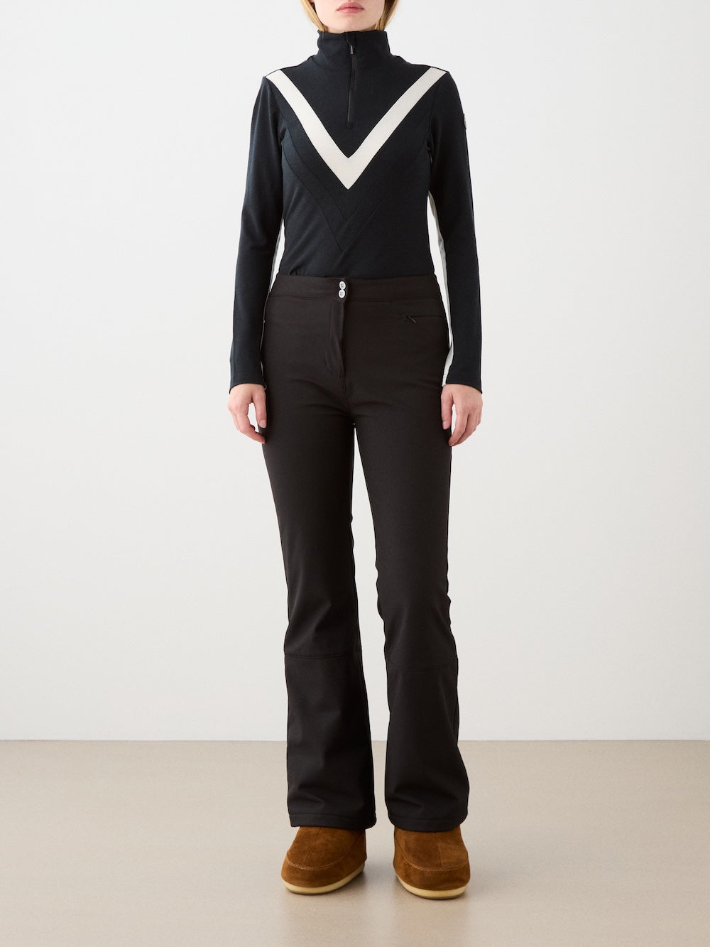 A person stands against a plain background wearing the We Norwegians Voss Chevron ZipUp Women, black flared pants, and brown suede boots. The top is black with a white V-shaped design. The person's face is not visible.