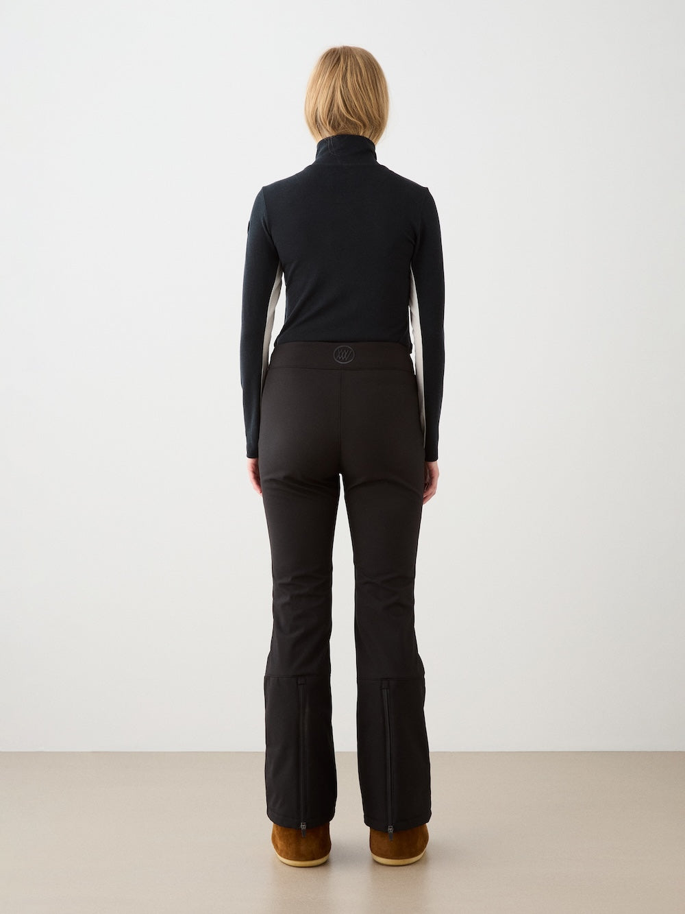 A person with blonde hair faces away, wearing the We Norwegians Voss Chevron ZipUp Women and fitted pants with brown boots, set against a plain light background.
