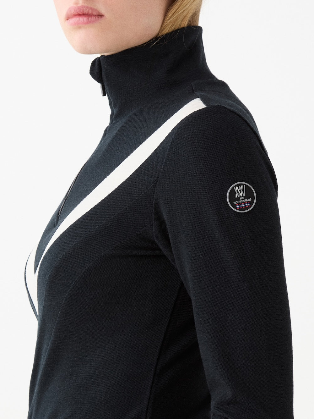 A woman wears the We Norwegians Voss Chevron ZipUp, a black Merino wool jacket with a white diagonal stripe and round logo patch on the sleeve, standing against a plain white background.