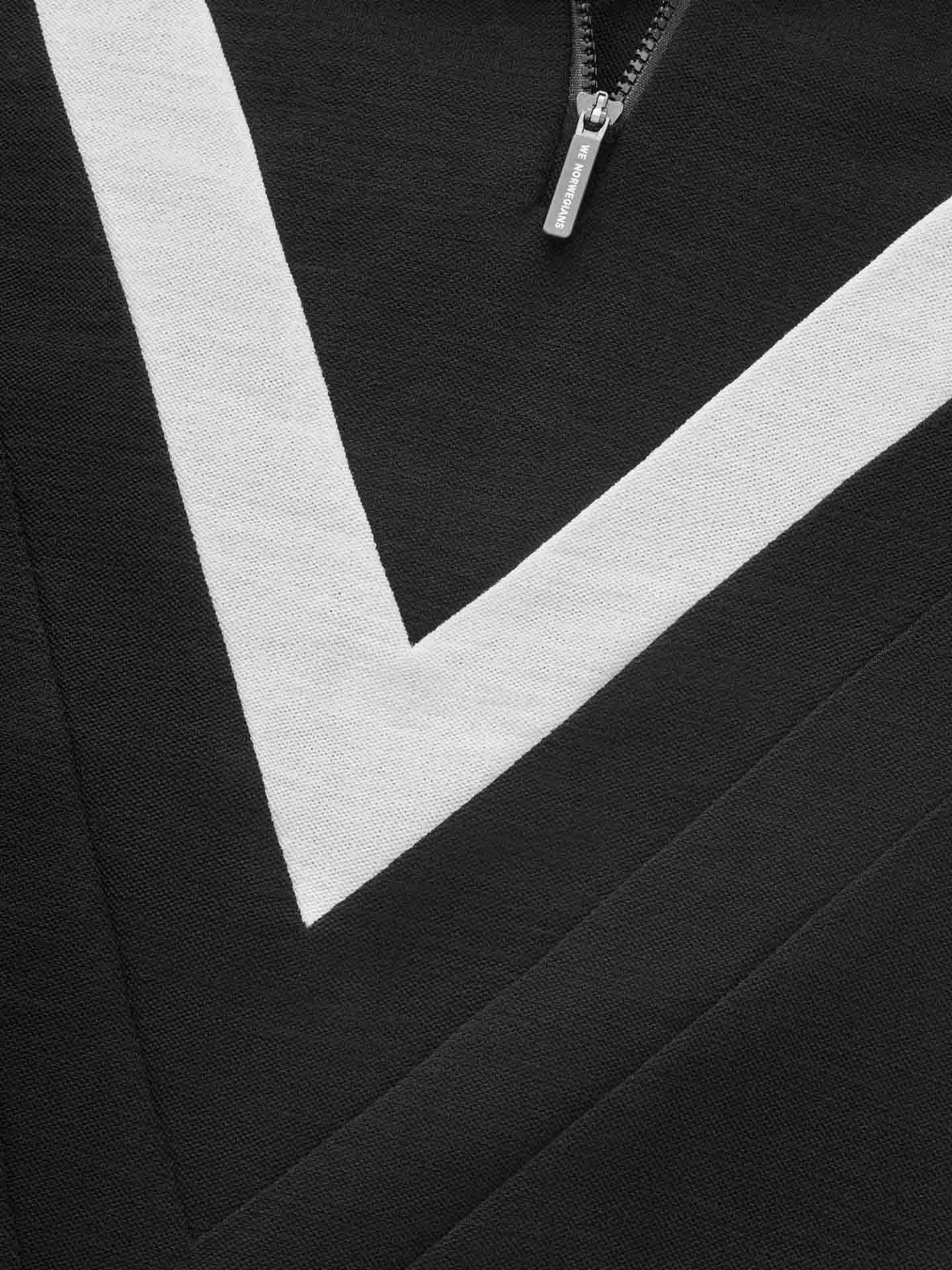 Close-up of the Voss Chevron ZipUp Women by We Norwegians, showcasing a black fabric with a white chevron pattern that evokes retro vibes. The partial view of the silver zipper pull engraved with "Y-3" draws attention, while the texture of the Super Fine Merino wool fabric and the sharp contrast between black and white colors are prominently featured.