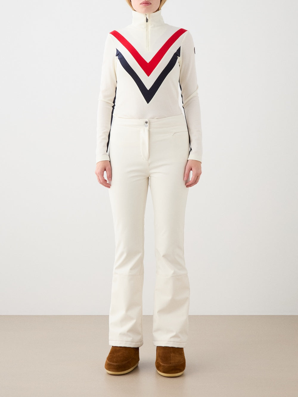 A person stands against a plain background wearing the Voss Chevron ZipUp Women by We Norwegians with cream pants and brown boots. This ski base layer blends warmth and style. Their hands are at their sides; their face is not visible.