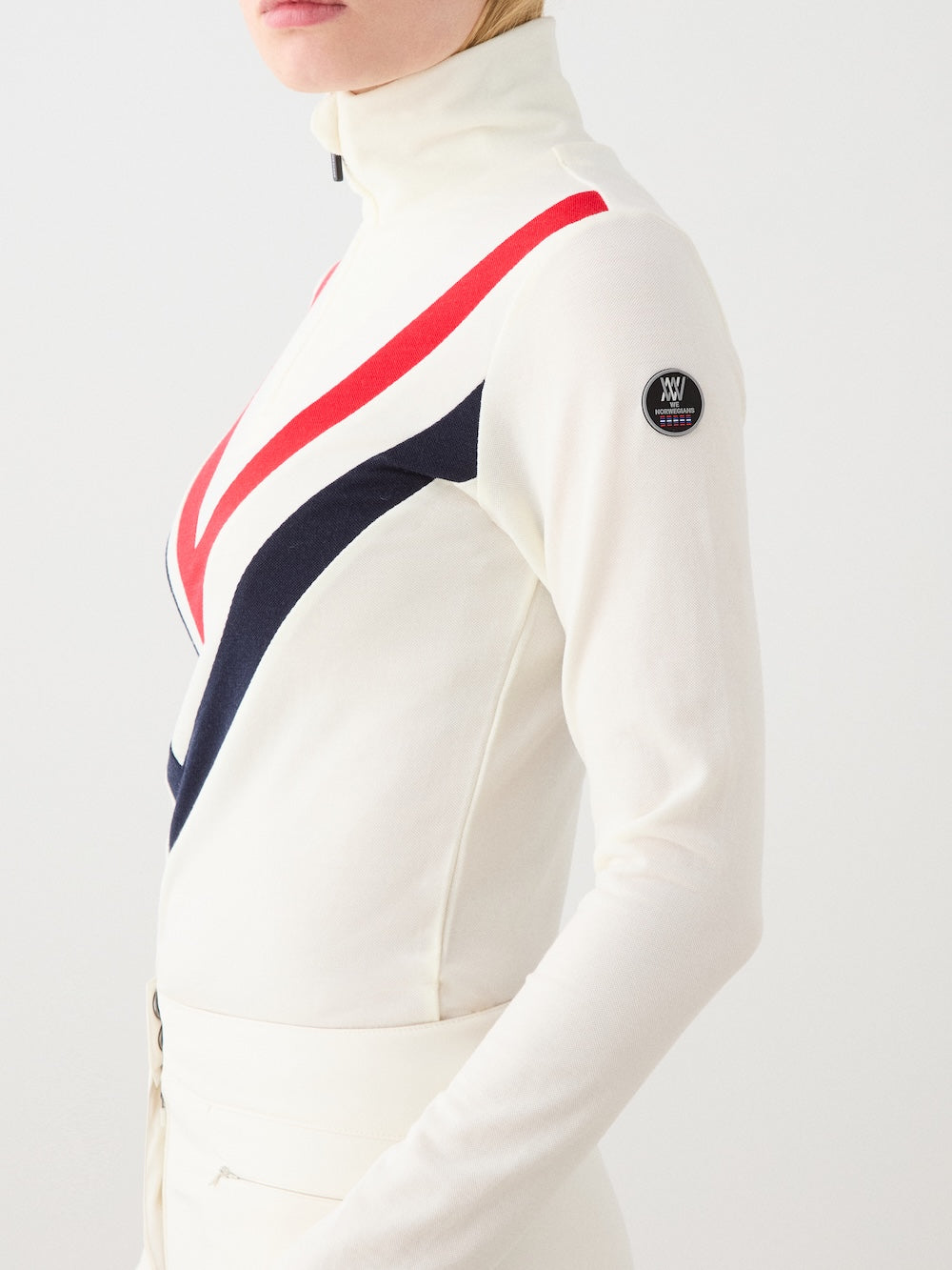 A woman stands against a white background wearing the We Norwegians Voss Chevron ZipUp, an off-white long-sleeve base layer with red and navy chevron stripes, a high collar, and a small black circular patch on the upper arm.