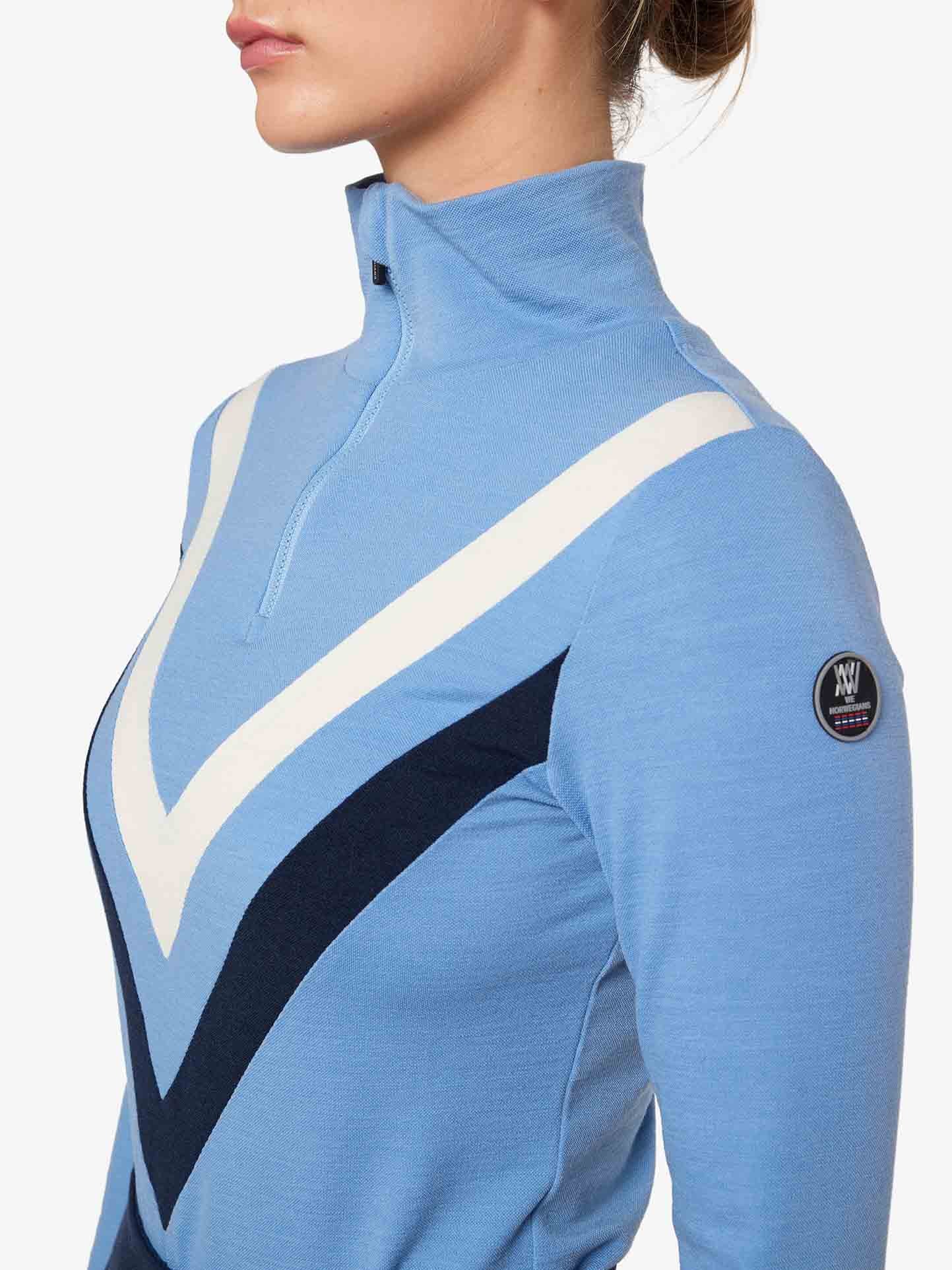 A person is wearing the Voss Chevron ZipUp Women by We Norwegians, featuring light blue Super Fine Merino wool with white and dark blue V-shaped patterns across the chest. The top has a half-zip collar and a small circular patch on the left sleeve, giving off retro vibes against a plain white background.