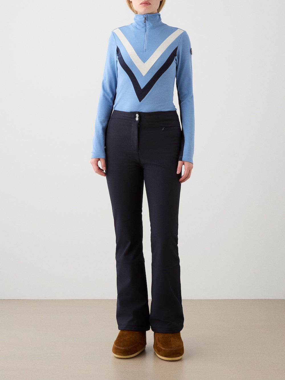 A person stands against a plain background wearing the We Norwegians Voss Chevron ZipUp Women in light blue with white and navy chevron stripes, paired with black flared pants and brown winter boots, hands relaxed at their sides.