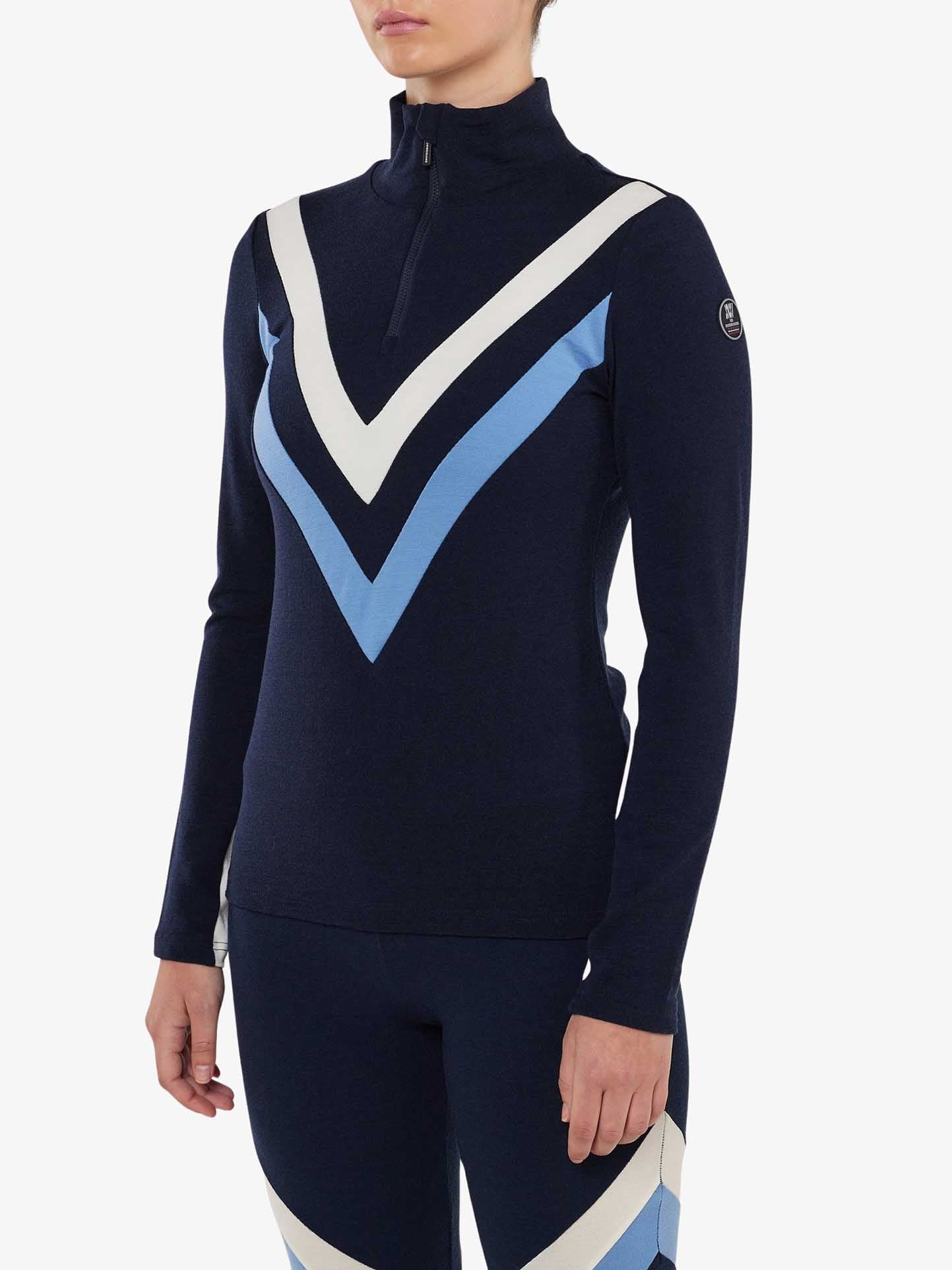 A person is wearing a Voss Chevron ZipUp Women by We Norwegians. The navy blue top features light blue and white V-shaped stripes on the upper and lower sections, crafted from Super Fine Merino wool with a half-zip collar. The photo does not show the person's face, evoking a retro aesthetic.