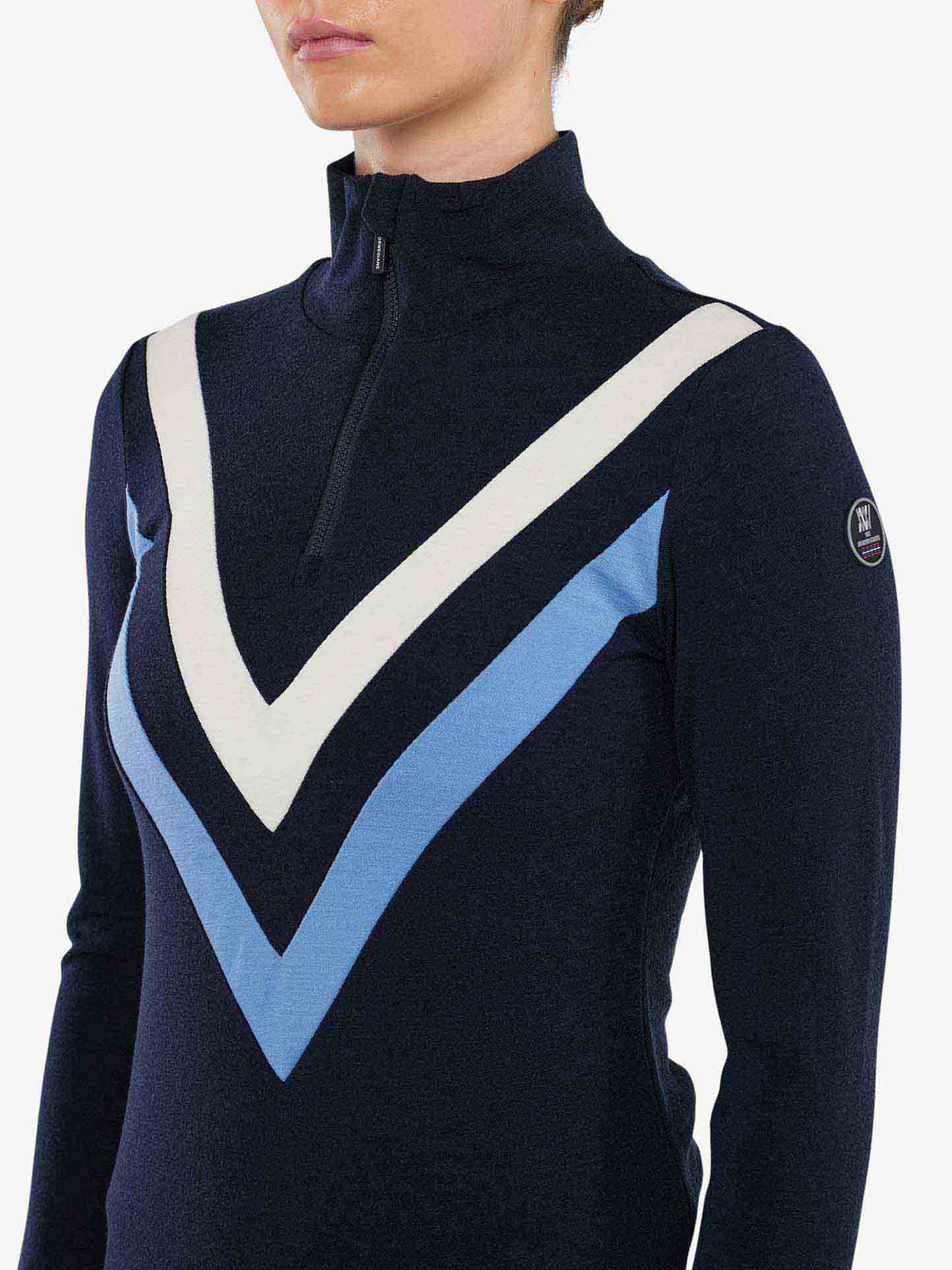 A person wearing a black We Norwegians Voss Chevron ZipUp Women sweater made of Super Fine Merino wool. The sweater features white and light blue V-shaped stripes across the chest, giving it retro vibes. The person's face is not fully visible, and there is a small logo on the left shoulder.