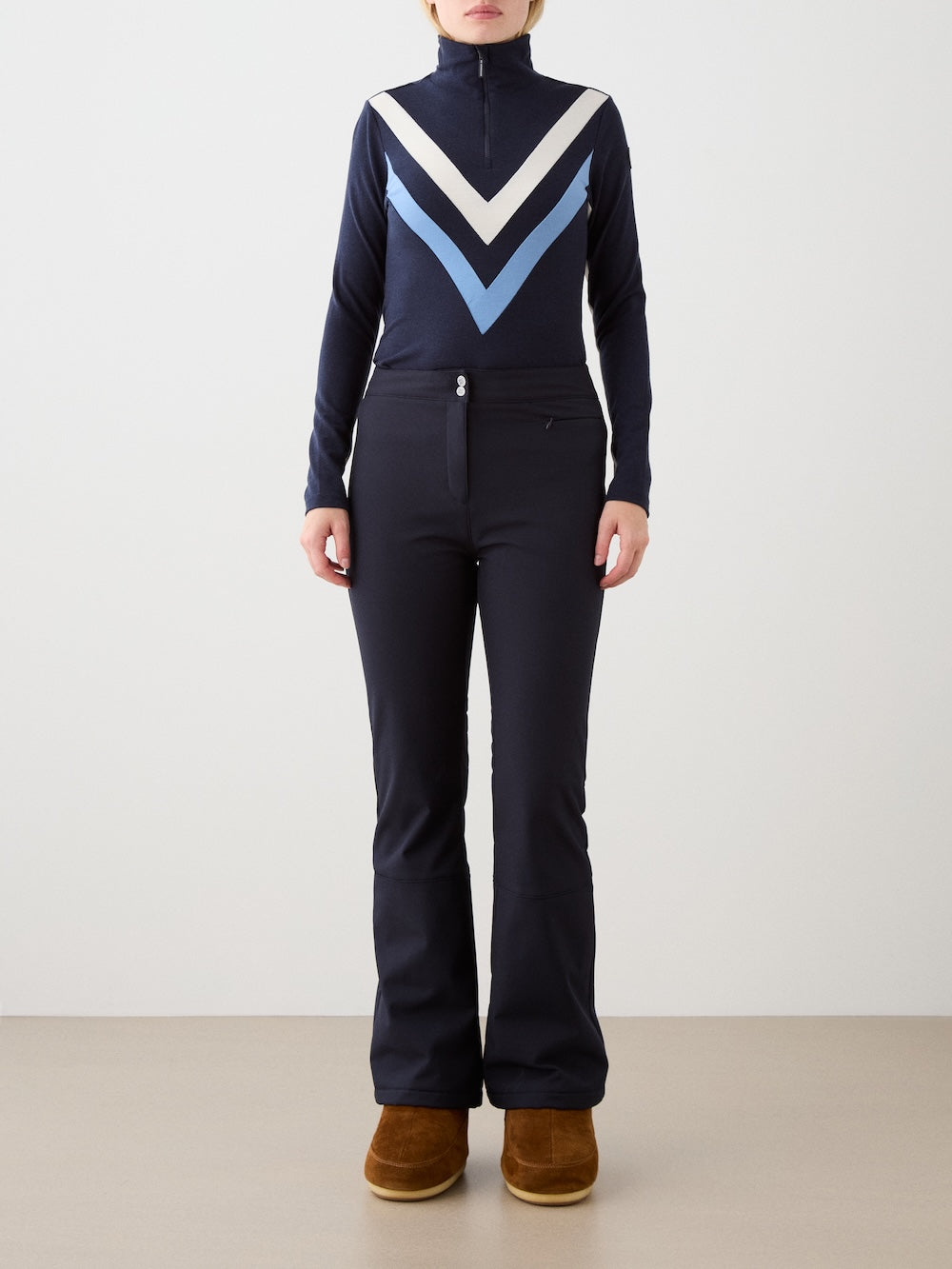 A person stands facing forward against a plain light background, wearing the We Norwegians Voss Chevron ZipUp Women in navy blue with white and light blue chevrons, fitted navy pants, and brown suede boots.