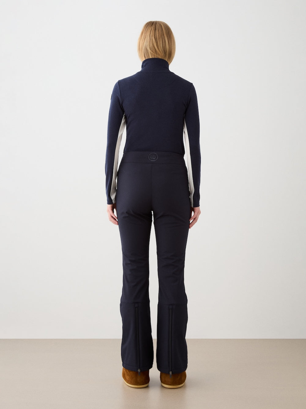 A person with short blond hair stands facing away, wearing the We Norwegians Voss Chevron ZipUp Women top, navy flared pants, and brown shoes, against a plain light background.