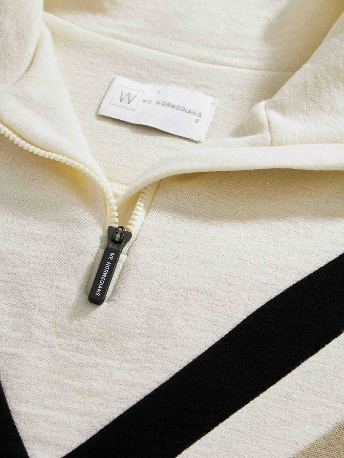 Close-up of an ivory-colored Voss Chevron ZipUp Women knit garment from We Norwegians, featuring a silver half zipper with "We Norwegians" engraved on the pull. Made from Super Fine Merino wool, it has a "We Norwegians" label sewn inside just above the zipper, while black triangular details on the lower part add subtle retro vibes.