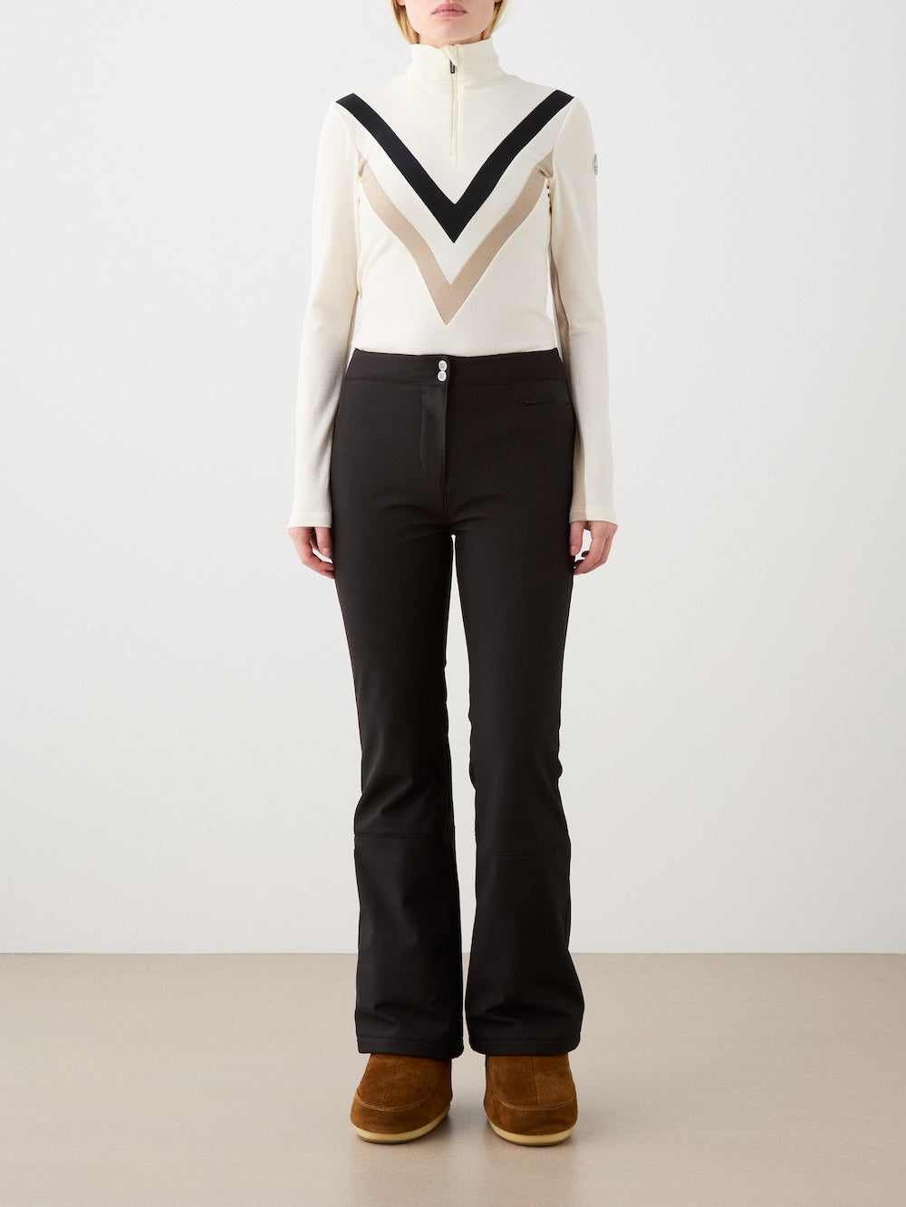A person stands against a plain background wearing the We Norwegians Voss Chevron ZipUp Women top with black fitted pants and brown boots.